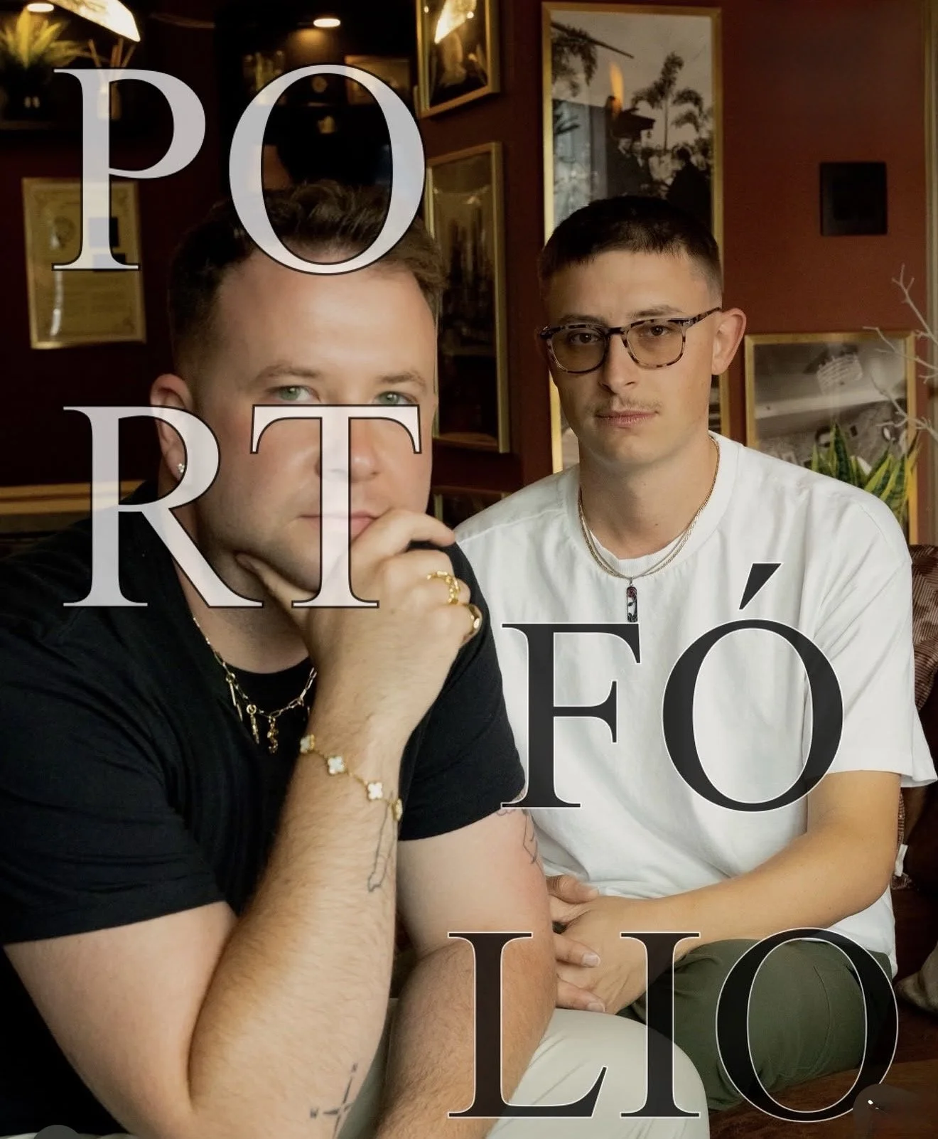 Two young men sitting at a table inside a restaurant or cafe, with framed pictures on the wall behind them. One man has short light hair and green eyes, wearing a black shirt and jewelry, resting his chin on his hand. The other man has short dark hair and glasses, wearing a white shirt. Overlaid text reads 'PO RTÉ ÉO LI O'.