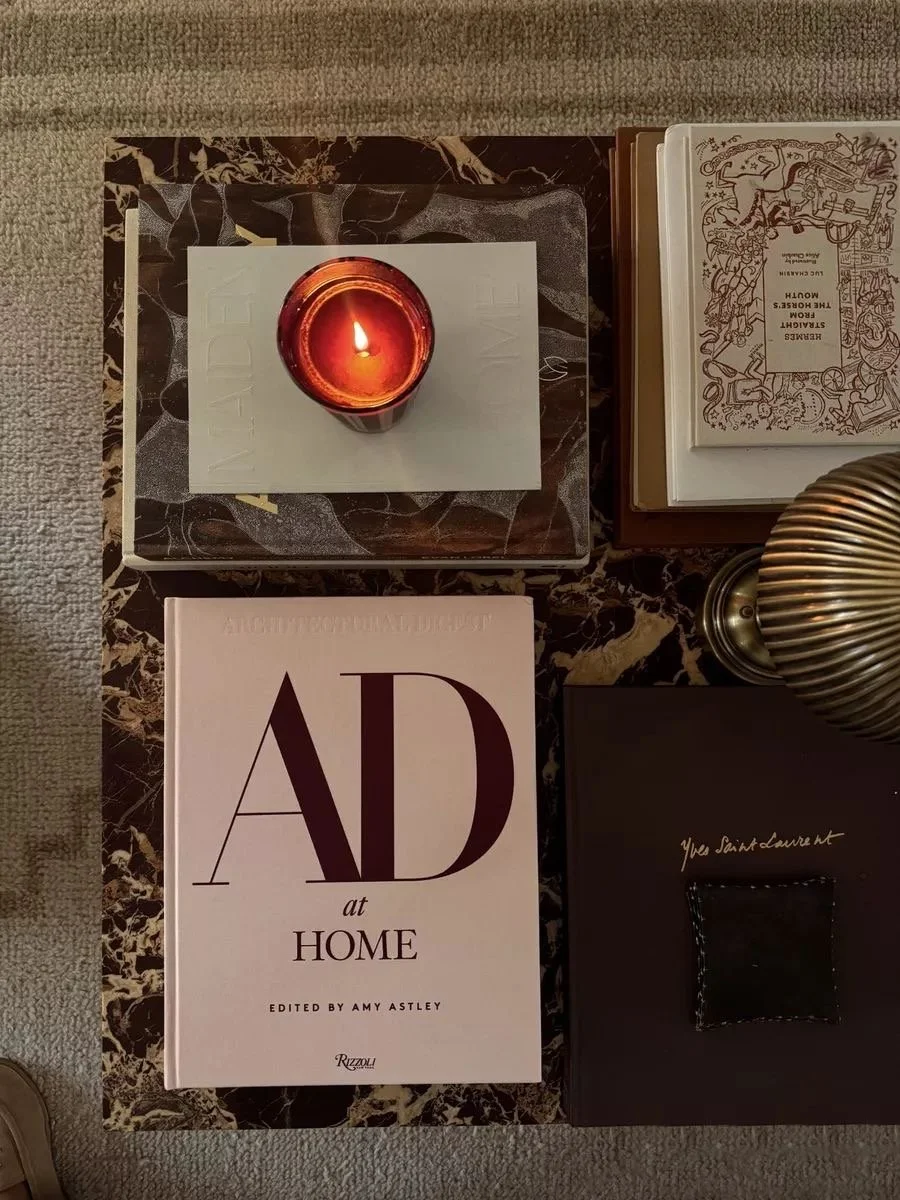 A candle in a small glass holder is on top of a white book. To the right are open books, one with intricate designs and another partially visible. Below the white book is a large black-and-white book titled 'AD at Home'. To the right is a metallic, ribbed lamp, and to the bottom right is a black box with a leather patch. The items are arranged on a dark marble surface with a beige carpet underneath.