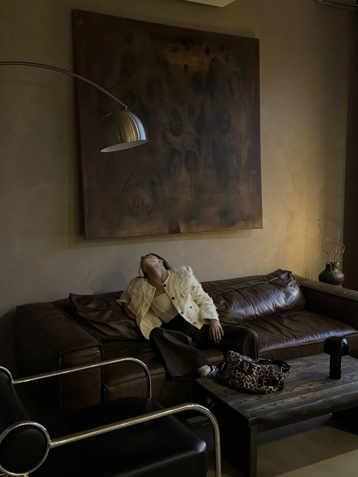 A woman sitting and leaning back on a dark brown leather sofa with her head tilted backwards and eyes closed, in a dimly lit room with beige walls, a large abstract painting and a potted plant on a side table.