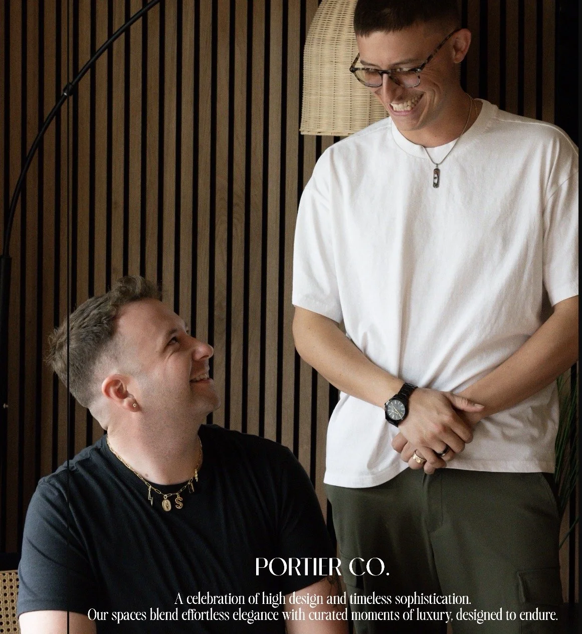 Two men smiling at each other inside a modern space with wooden wall panels; one wearing a black shirt with jewelry, the other in a white t-shirt with glasses and a watch, standing near a lamp.