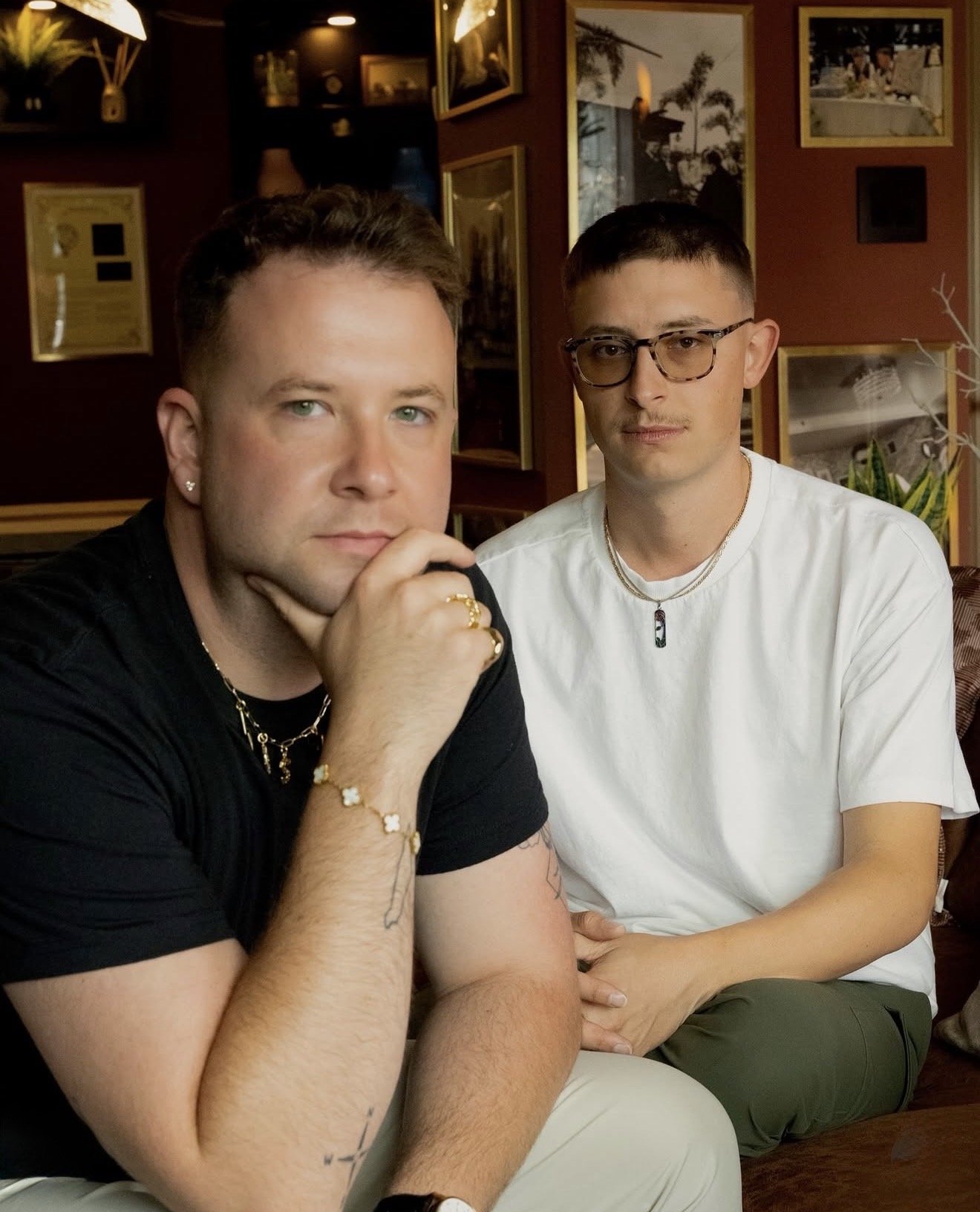 Two young men sitting close together in a cozy room with framed pictures on the wall behind them. The man on the left has light green eyes, short brown hair, and is wearing a black shirt, jewelry, and a bracelet. He has his hand on his chin. The man on the right has glasses, short brown hair, and is wearing a white T-shirt and layered necklaces. They are looking directly at the camera.