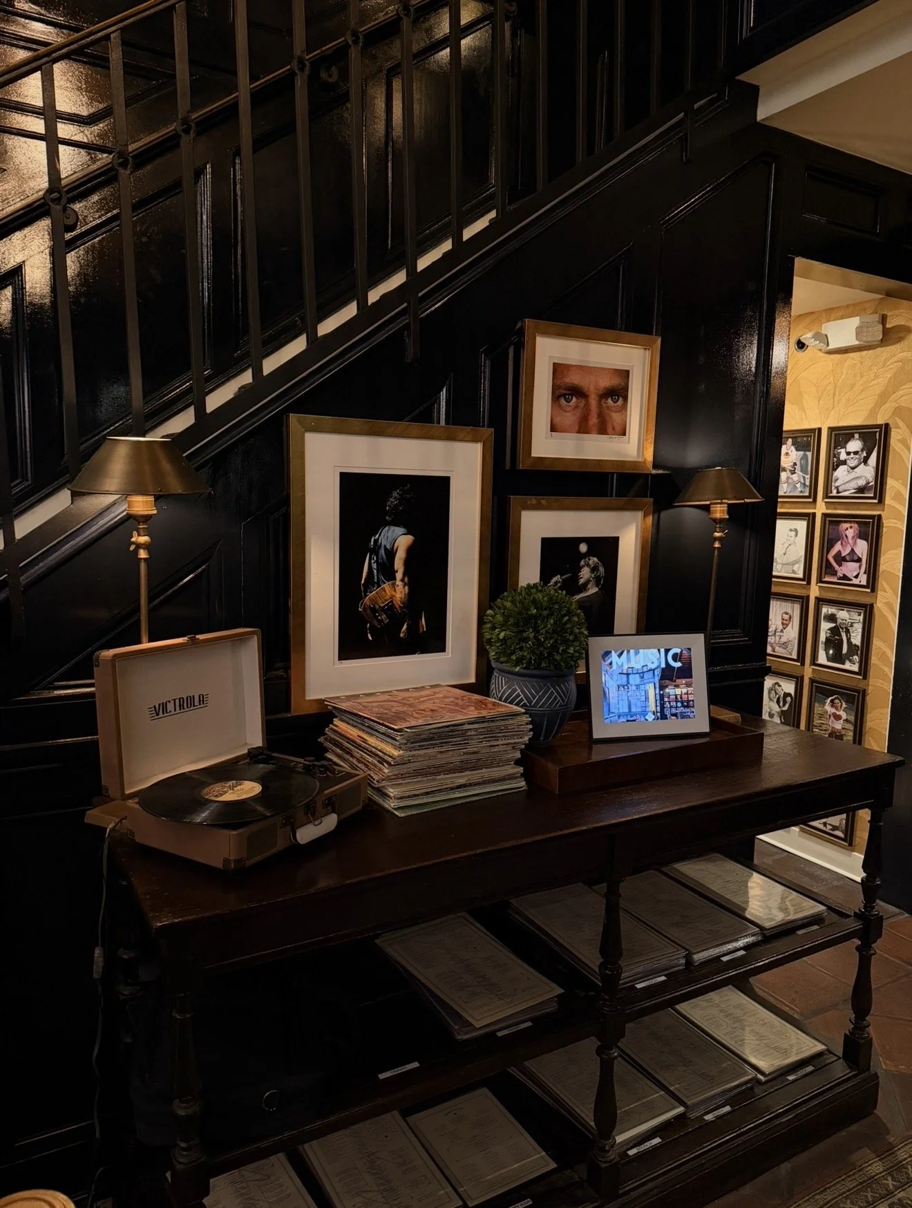 Interior view of a room with black paneled walls, a staircase with black railing, and framed photos of various people on the walls. There is a desk with a turntable, stacks of vinyl records, a small digital photo frame, and two table lamps.