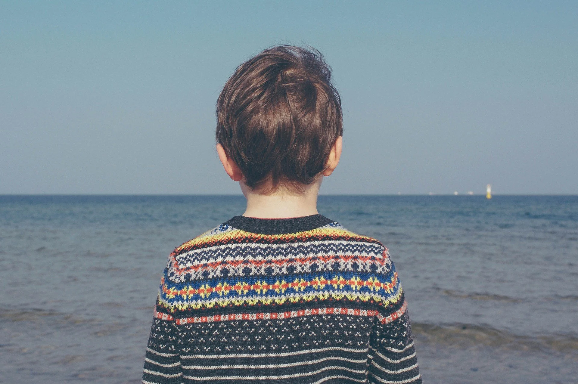 Child in a patterned sweater standing on a beach and looking out toward the ocean