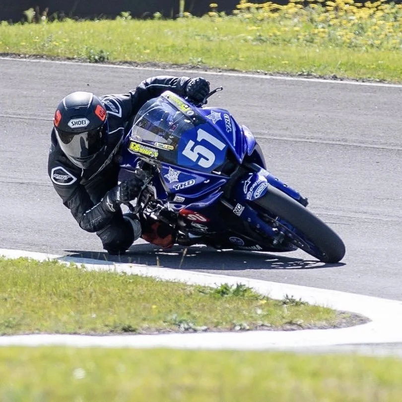 Motorcycle racer leaning into a turn on a race track, wearing black racing suit and helmet, with a blue motorcycle numbered 51.