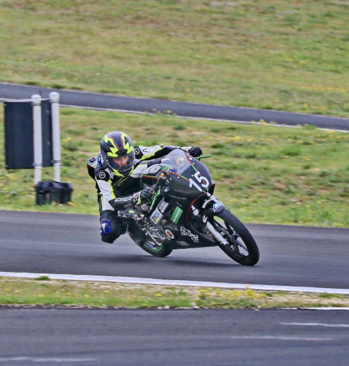 A motorcyclist wearing a black and yellow helmet and suit leans into a turn on a racetrack, riding a black race bike with the number 15.