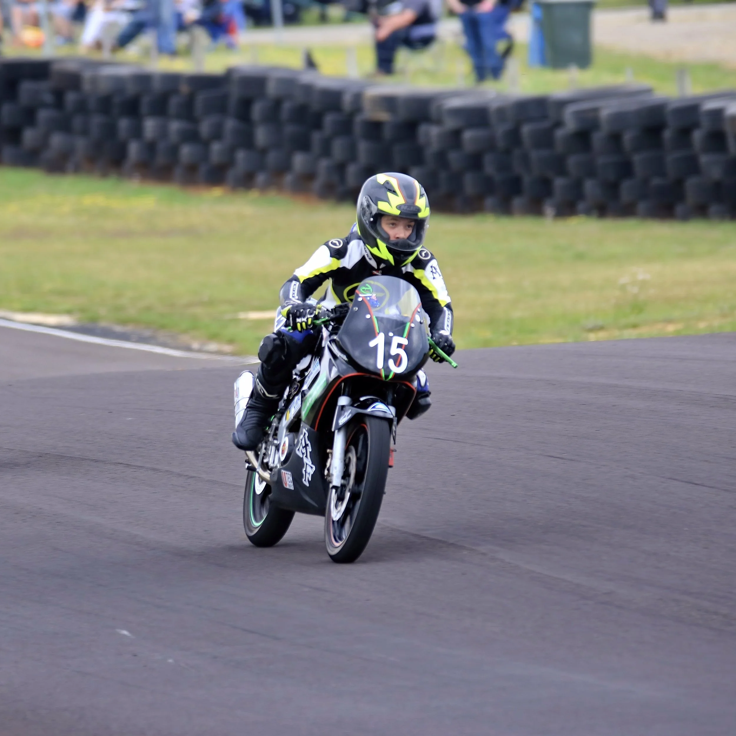 A motorcycle racer wearing a black and yellow helmet and racing suit riding on a racetrack, with tires stacked in the background and spectators in the distance.