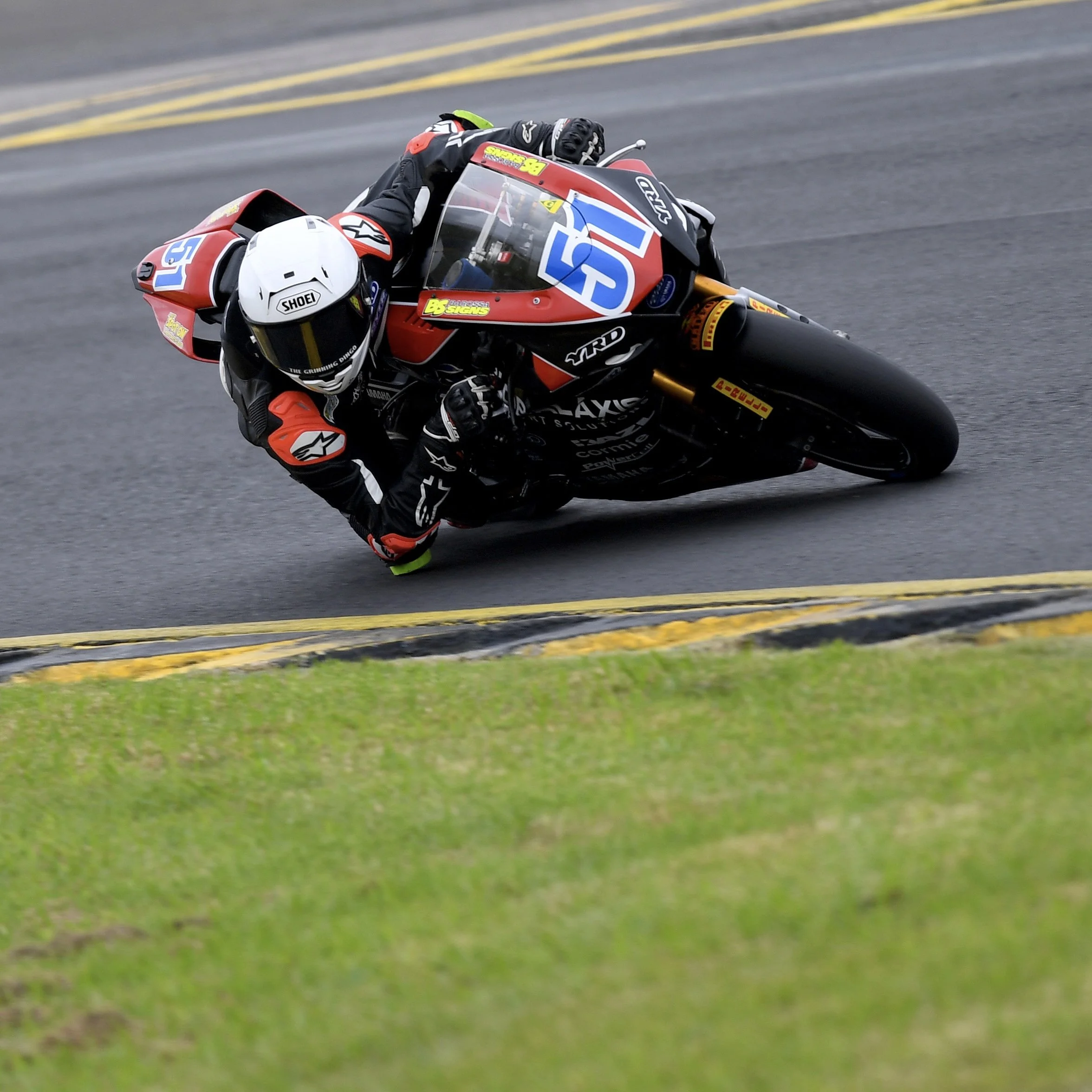 A motorcycle racer in black and red racing suit leaning into a turn on a racetrack, wearing a white Shoei helmet, with a yellow and black barrier wall in the background and green grass on the track's side.