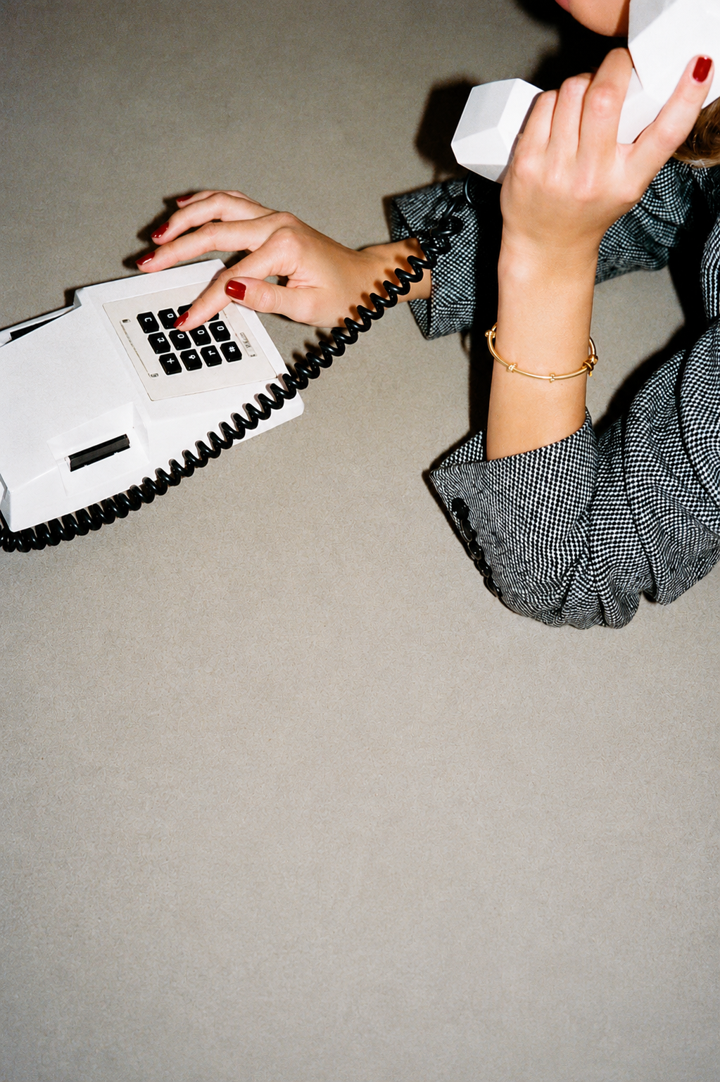 A woman using a white rotary phone with a coiled cord, wearing a checkered blazer, red nail polish, and a gold bracelet, seated at a beige table.