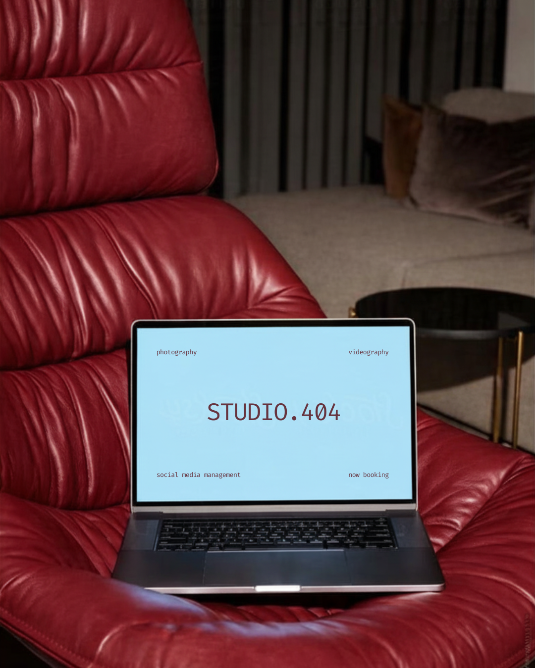 A laptop on a red leather chair with the screen displaying 'STUDIO.404' and small text that reads 'photography', 'videography', 'social media management', and 'now booking'.