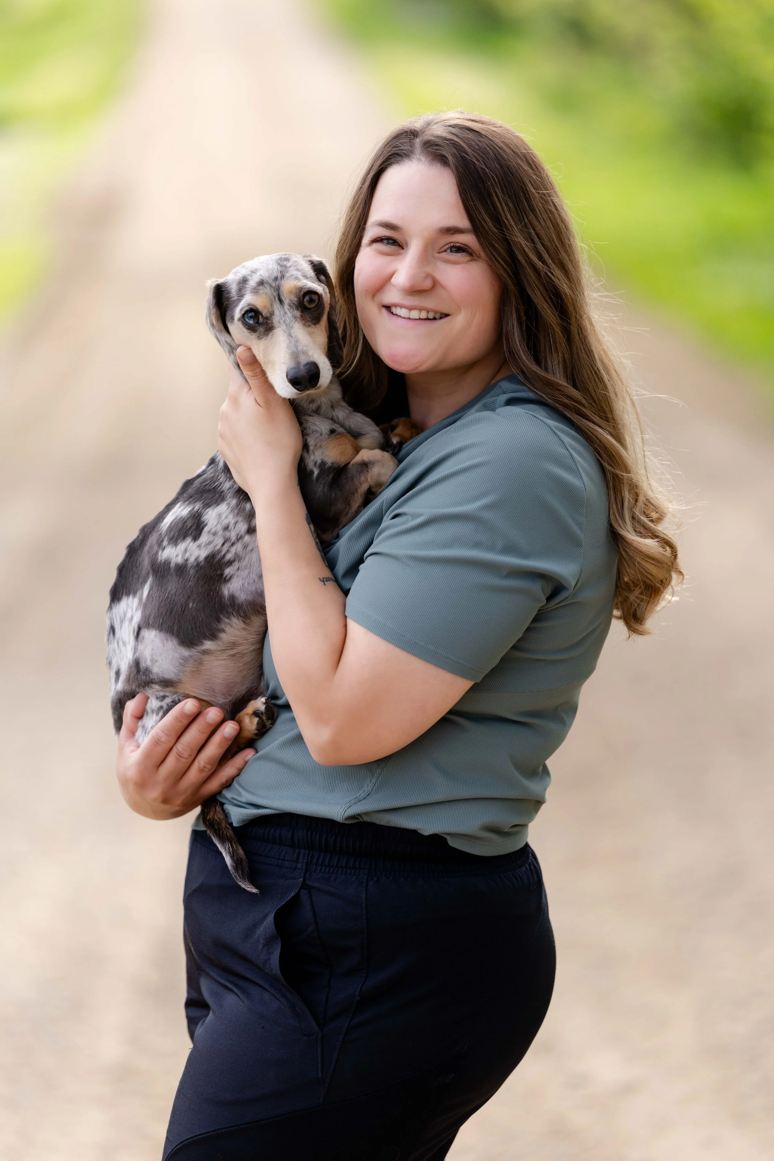 A woman in a gray shirt smiling while holding a dappled gray and black dog outdoors with a blurred green nature background.
