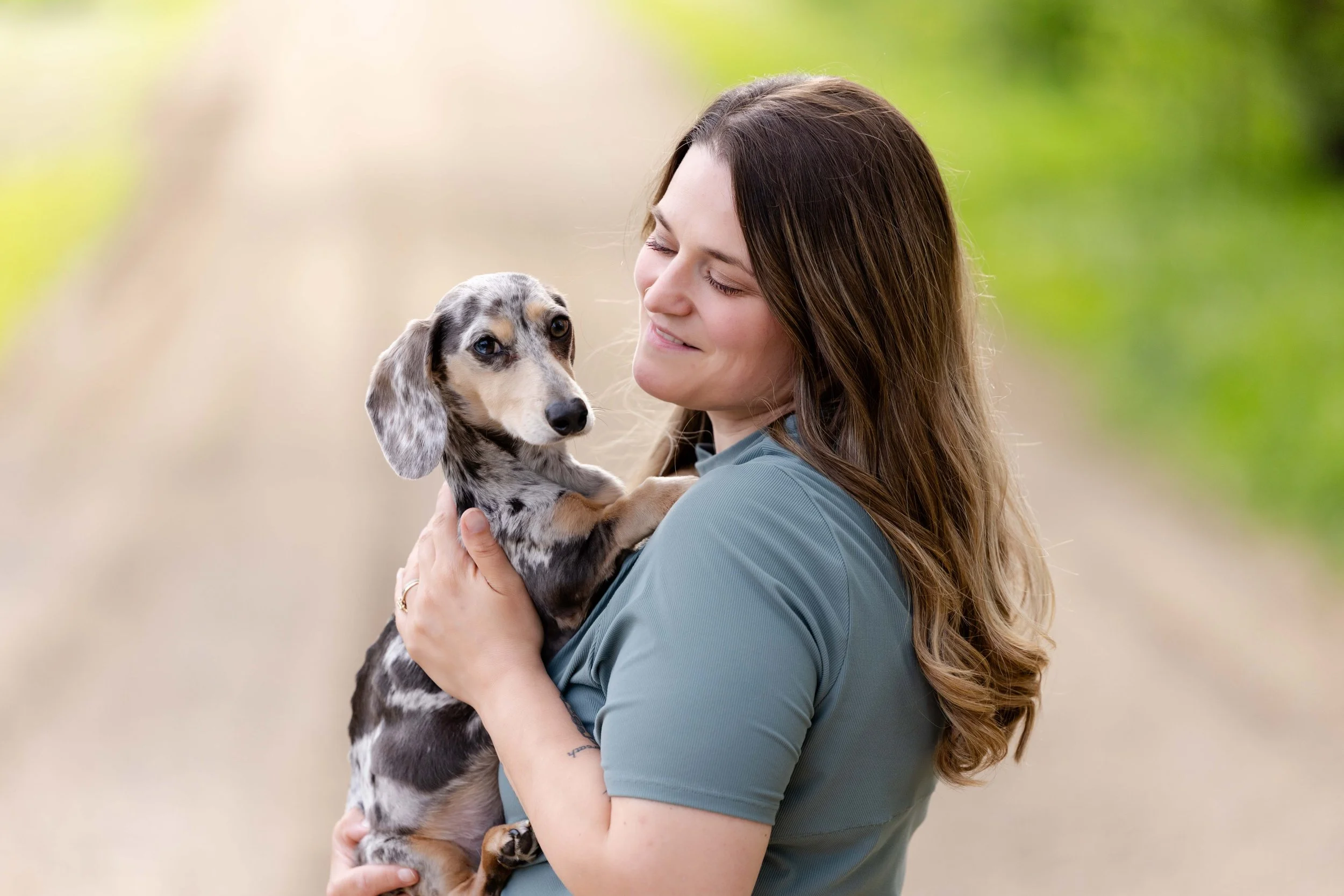 A woman with long brown hair holding a young red, gray, and black dappled Dachshund puppy with floppy ears in her arms outdoors, with a green blurred background.