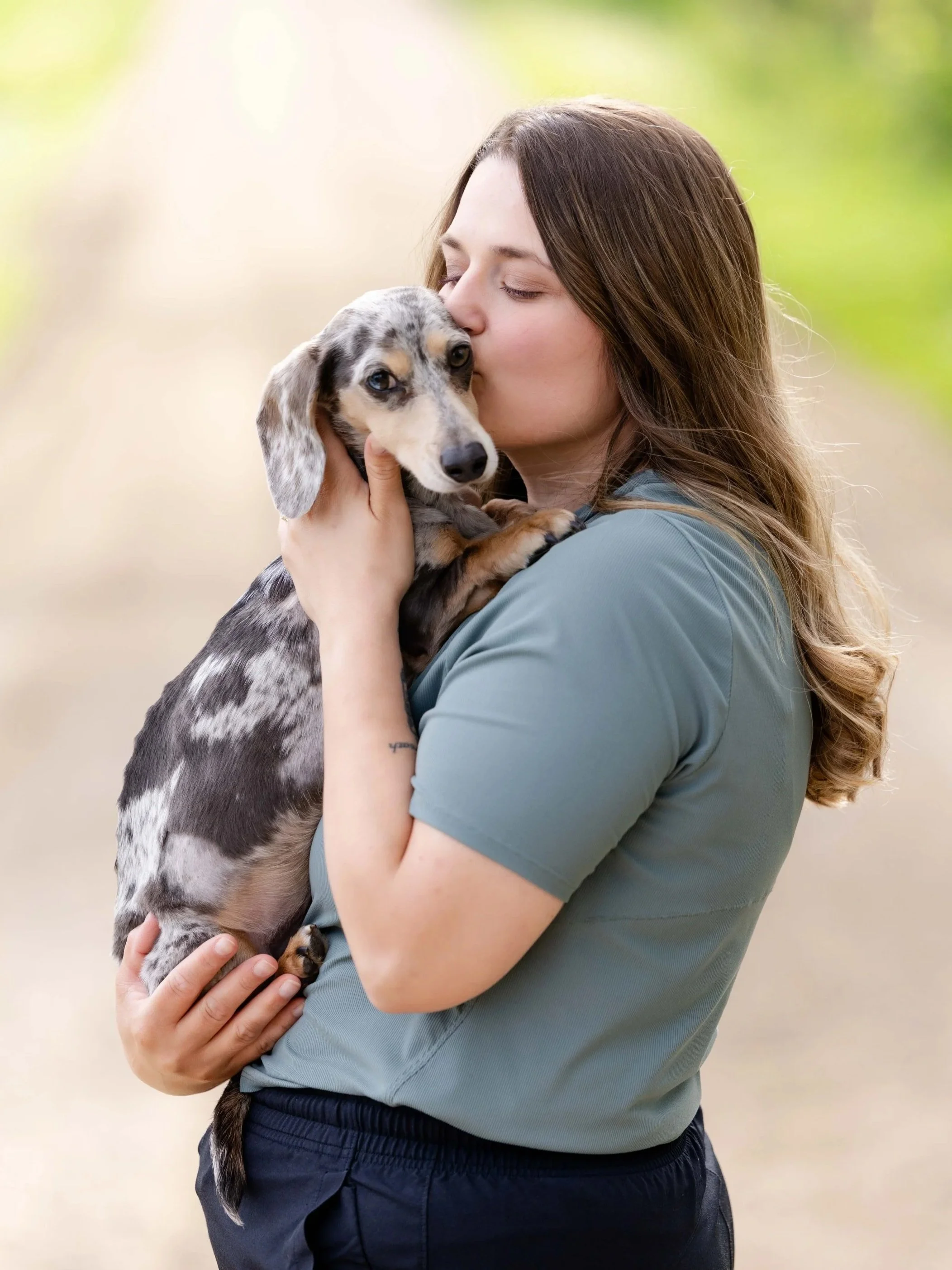 A woman with long brown hair and a gray shirt holding and kissing a spotted puppy with floppy ears in her arms outside with a blurred green background.