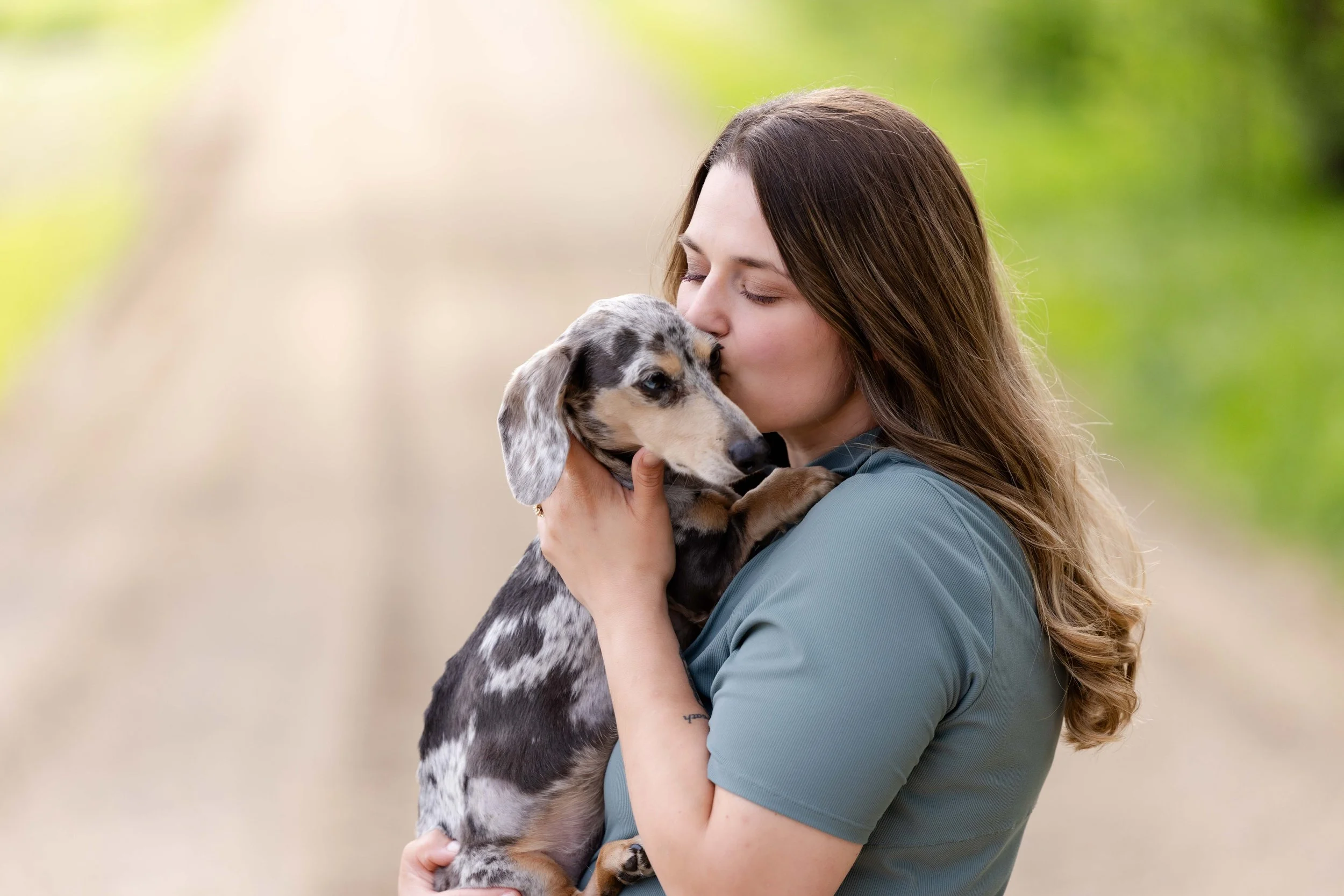 A woman with long brown hair holding and kissing a merle-patterned dachshund puppy outdoors with a blurred green background.