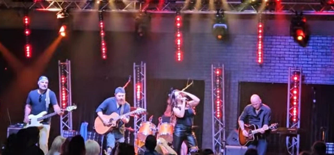 A live musical performance on stage with a female singer and three male guitarists, stage lighting, and audience in front.