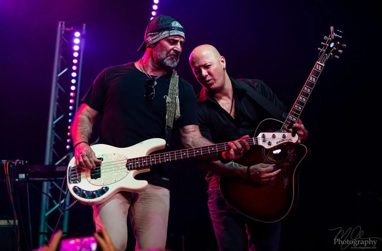 Two male musicians playing guitars on stage under purple and blue stage lights.