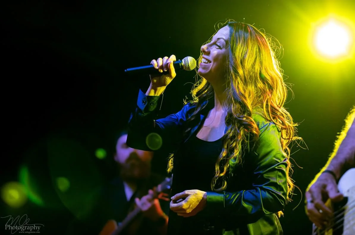 A woman singing into a microphone during a live music performance, with colorful stage lighting. Sarah Moon as Alanis Morissette, performing at an Infatuation Junkie Live Performance.
