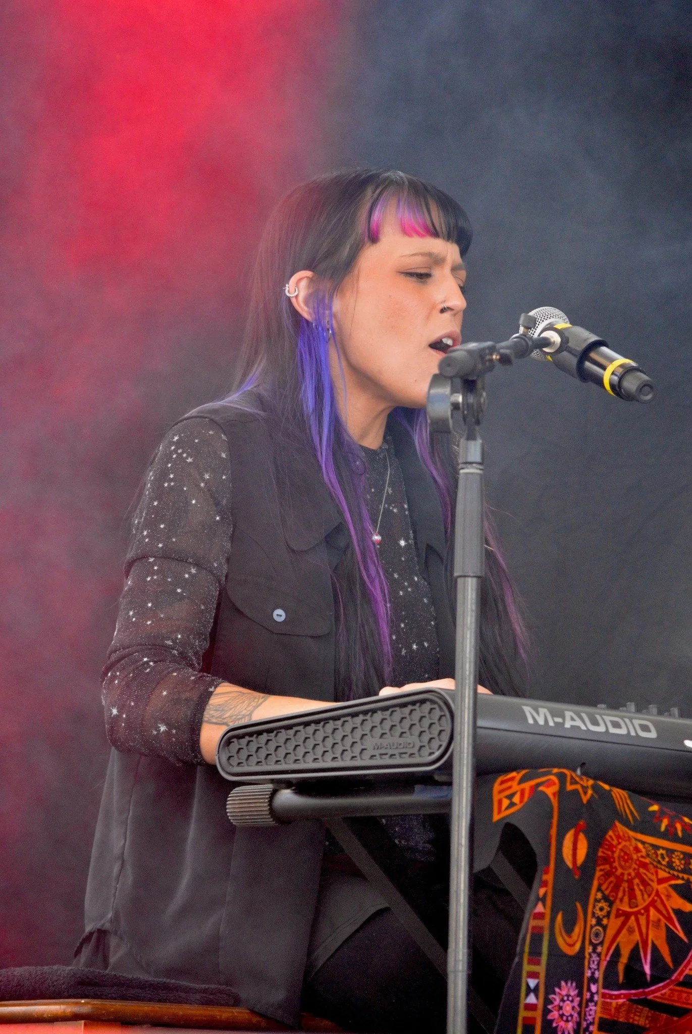 A woman with purple and pink hair singing or speaking into a microphone on a stage, with a colorful patterned fabric in the foreground and fog or haze in the background.