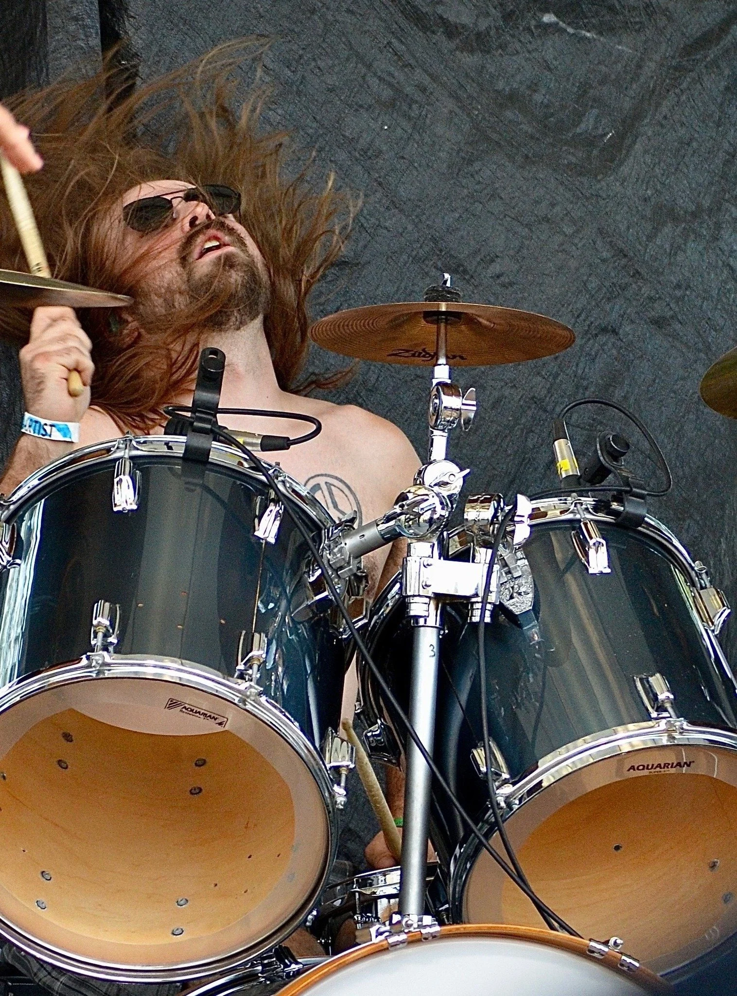 A man with long hair, beard, and sunglasses lying on the ground with his head near a drum set, playing drums.