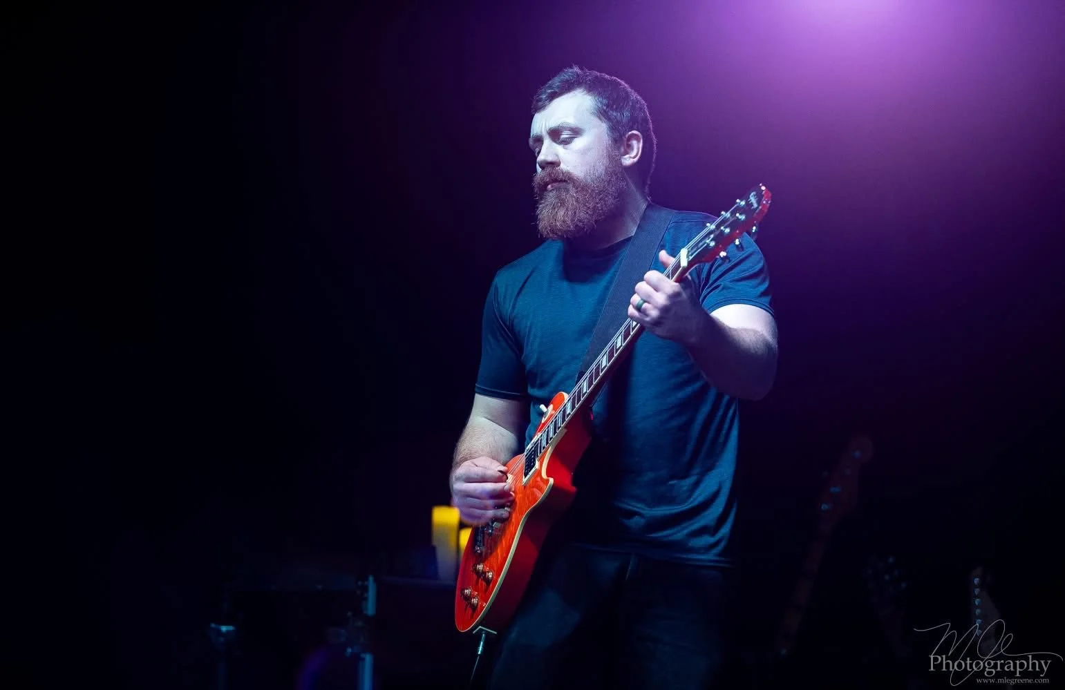 A bearded man in a dark blue t-shirt playing an orange electric guitar on stage with purple lighting.