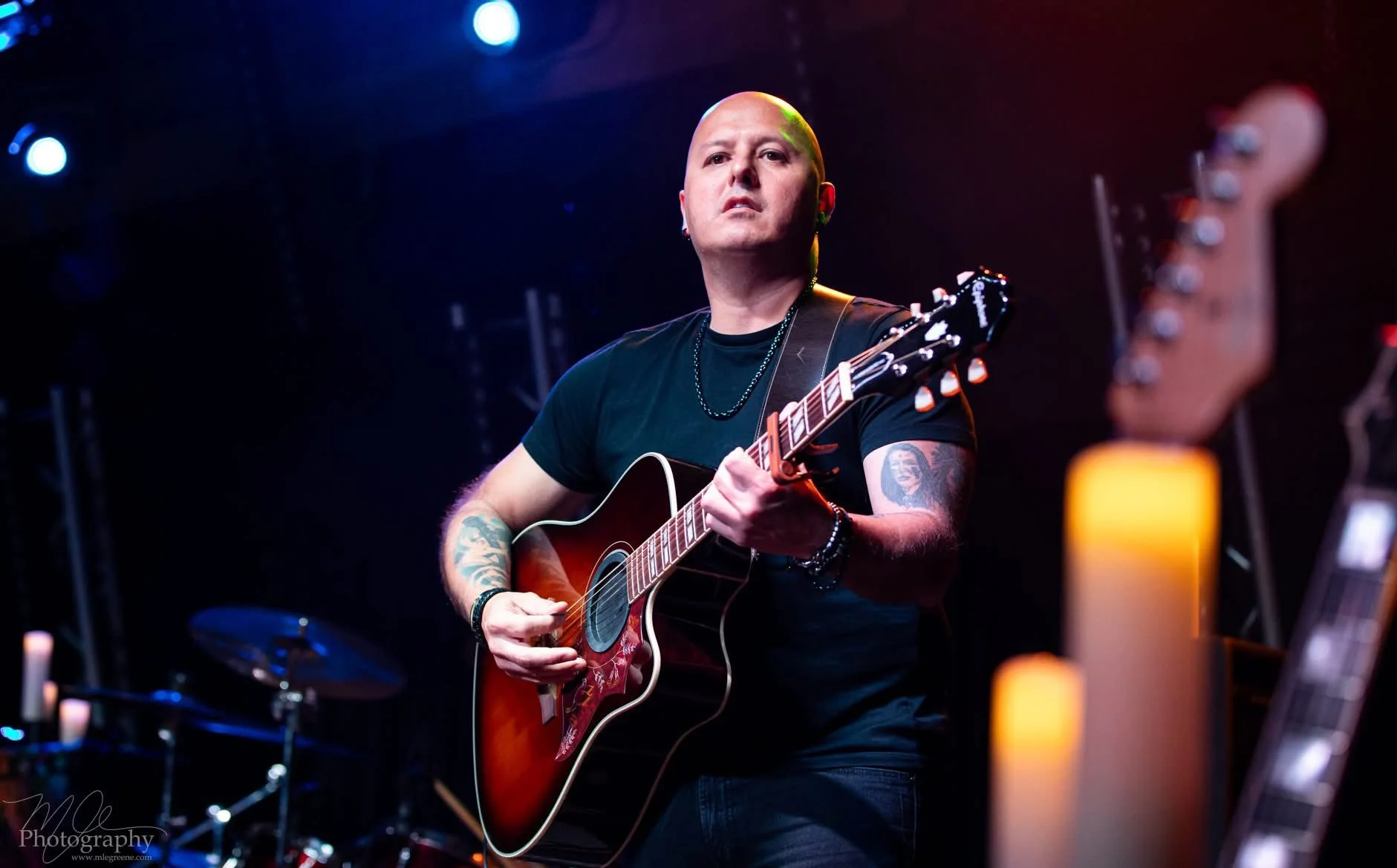A bald man playing an acoustic guitar on stage, illuminated by stage lights, with blurry background drums and other musical equipment.