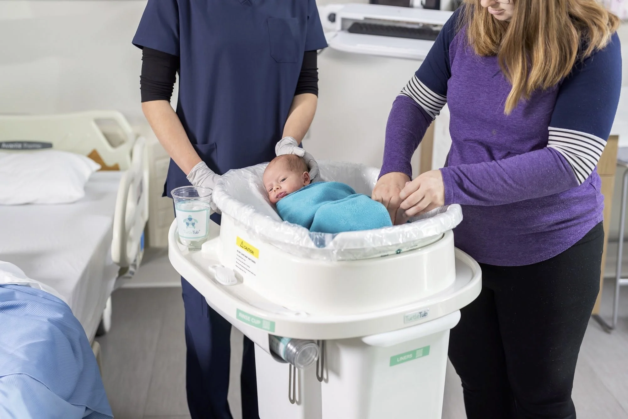 A nurse and a woman tending to a newborn baby in a hospital with baby in the TurtleTub swaddle bathing cart.