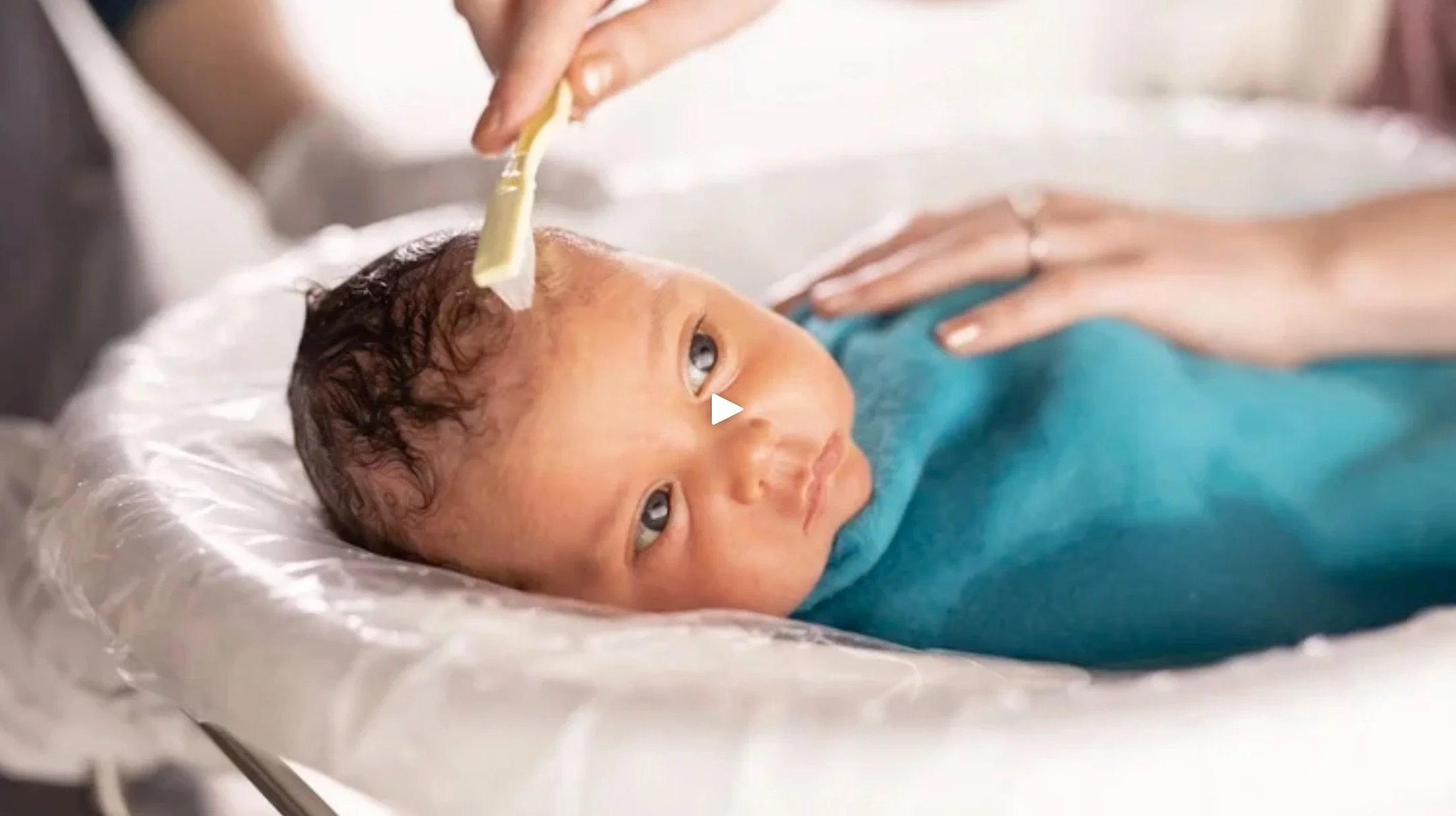 A newborn baby with curly dark hair getting a swaddle bath in the TurtleTub.