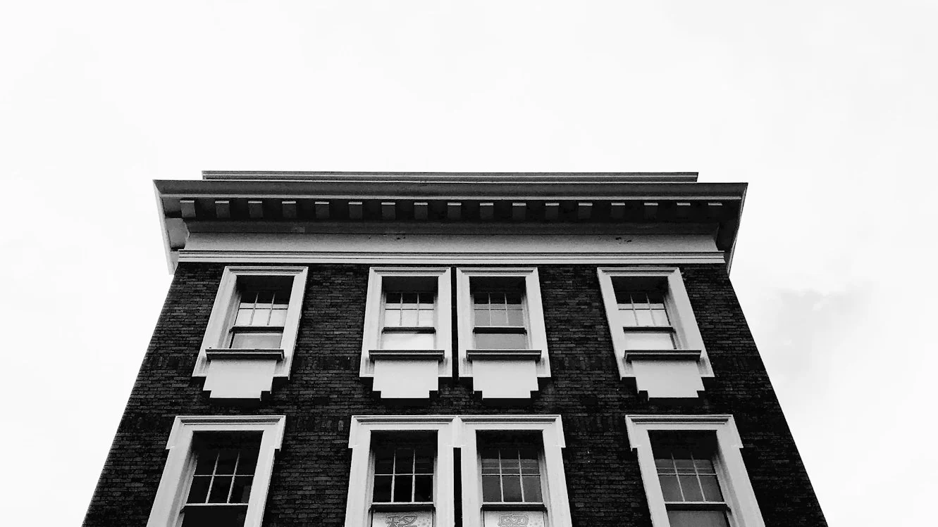 Black and white photo of the upper floors of a brick building with decorative window trims, viewed from below.