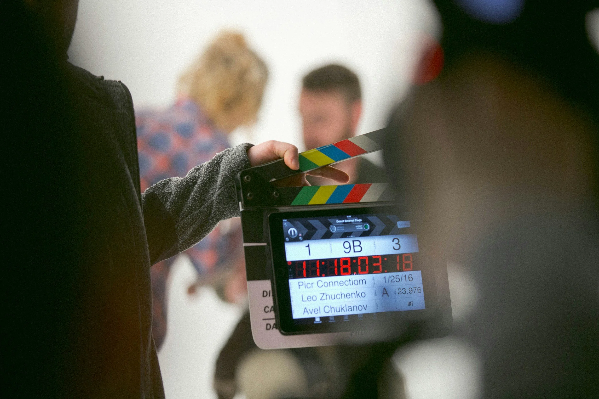 Close-up of a person holding a clapperboard in front of a monitor displaying scene information, with blurred background of two people.