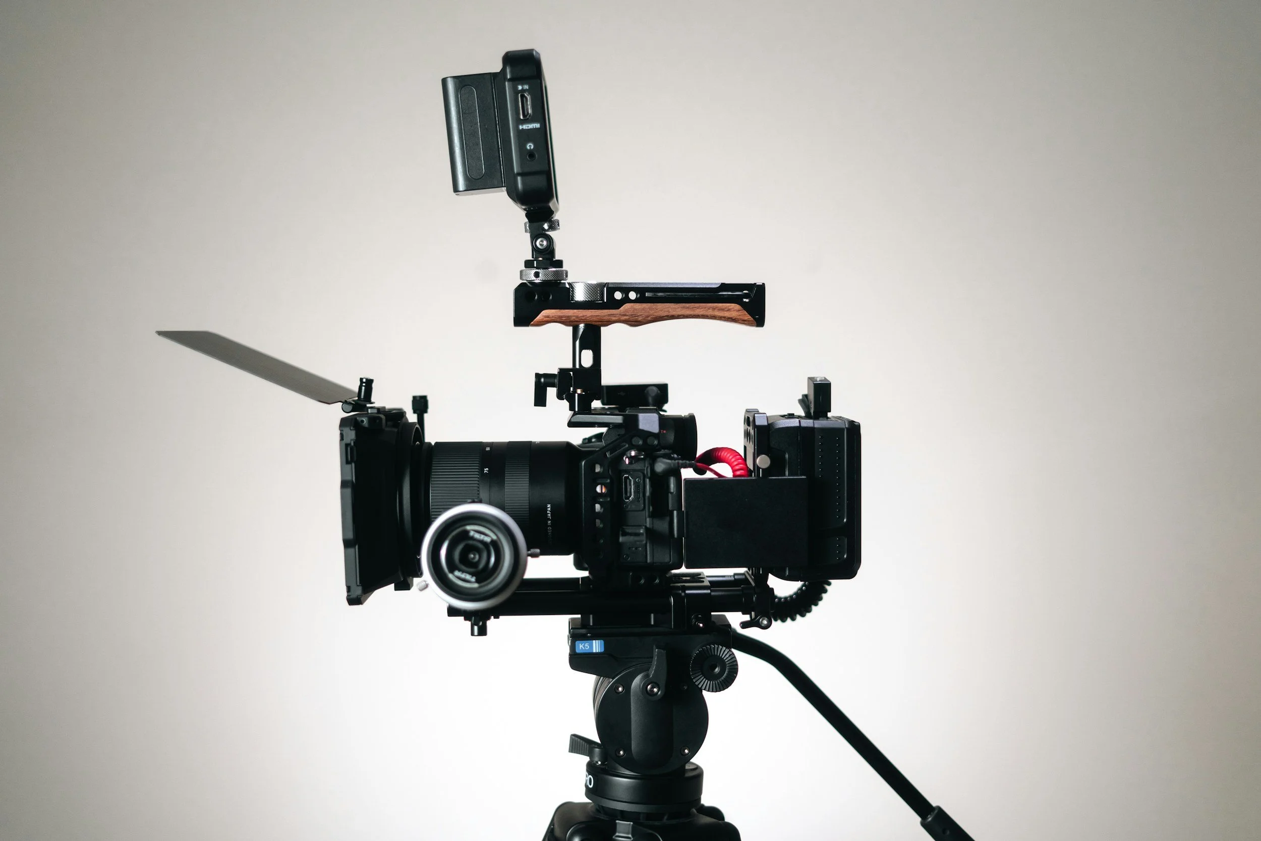 A professional camera mounted on a tripod with a monitor, microphone, and other camera equipment attached, set against a plain light gray background.