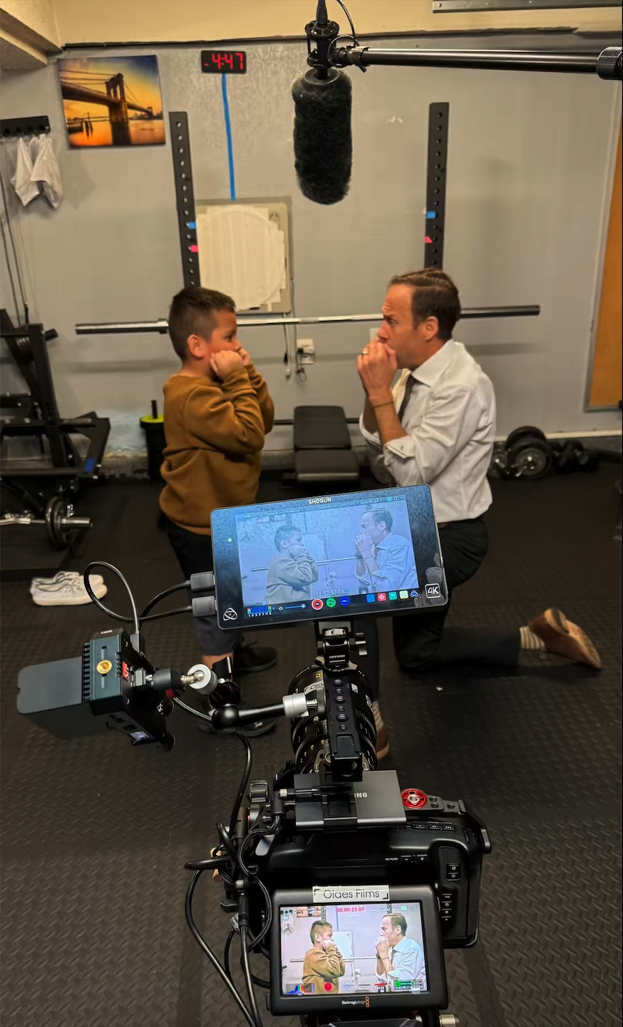A young boy and an adult man are facing each other in a gym, holding their hands near their faces in a playful imitation of a boxing pose while smiling. A camera on a stabilizer records their interaction, and the camera's screen shows a live view of the scene.