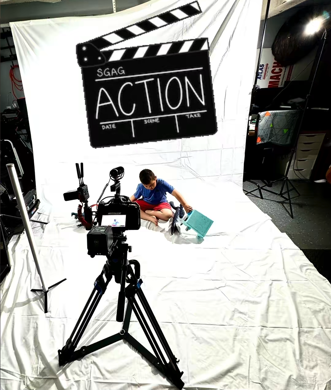Child sitting on a white sheet in a film studio, surrounded by lighting and camera equipment, with a clapperboard sign that says 'ACTION' hanging above.