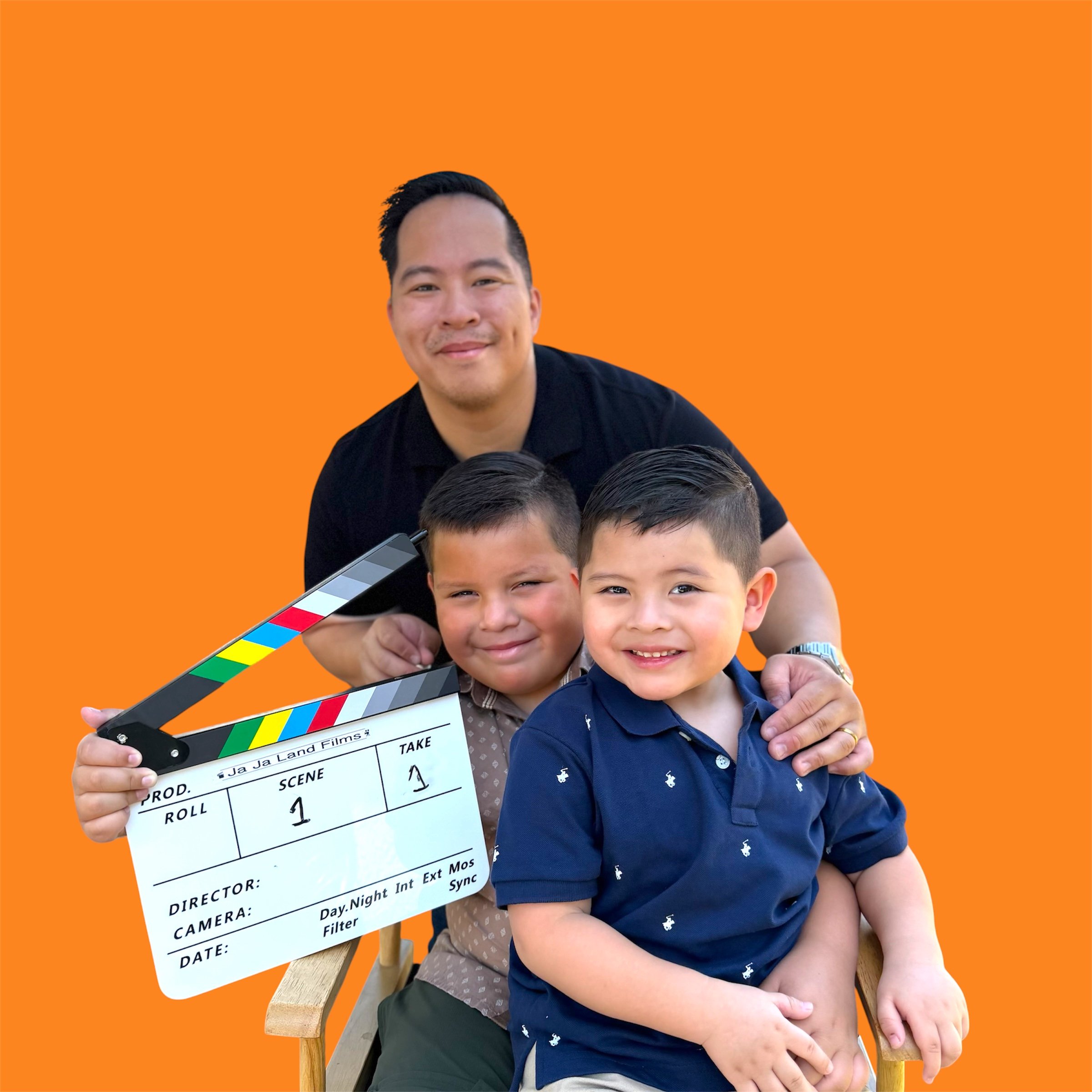 A smiling man sitting on a wooden chair with two children in front of him, holding a film slate, on an orange background.
