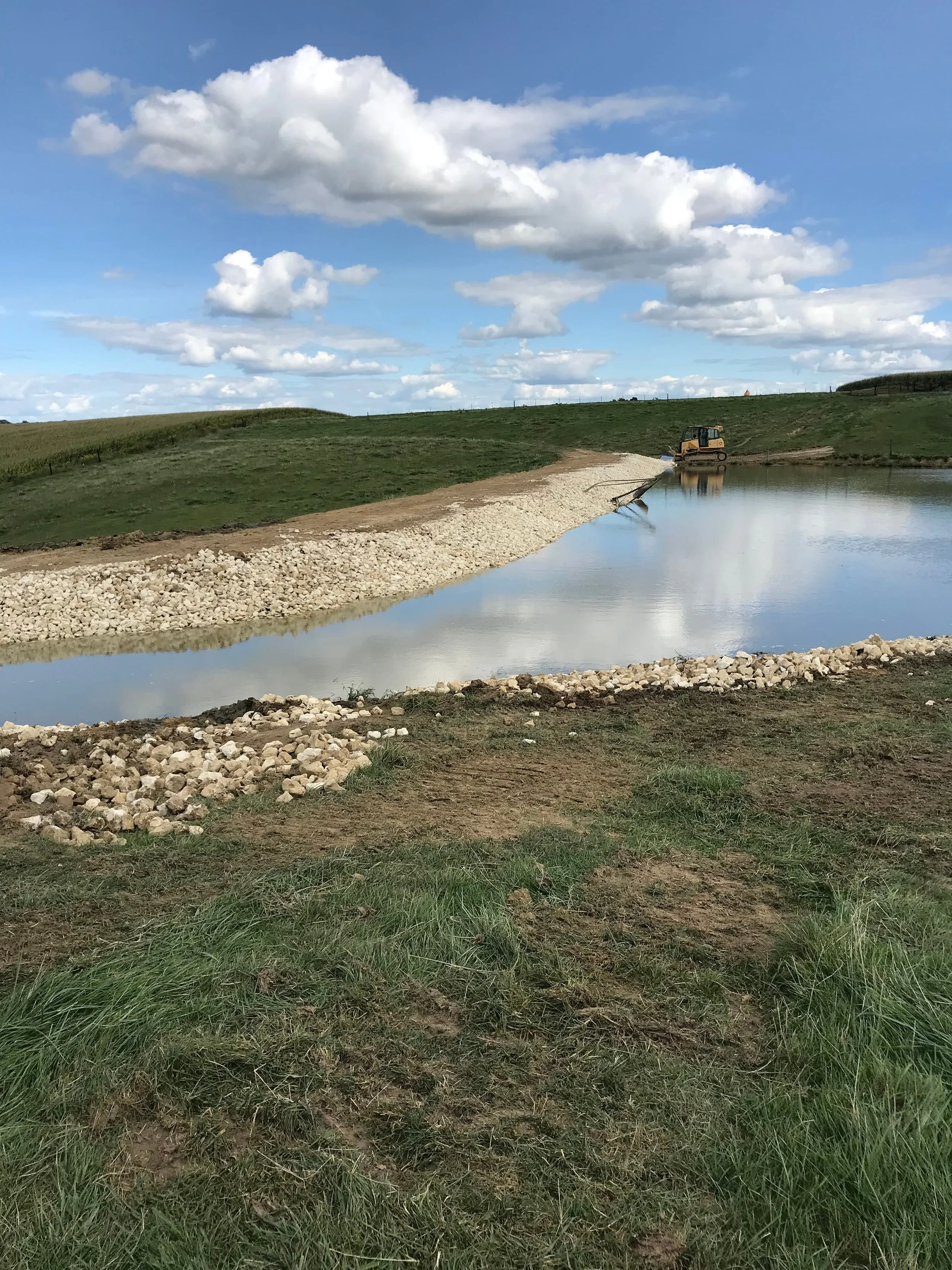 A construction worker operating a bulldozer on a small body of water with a grassy hill and blue sky with clouds in the background.