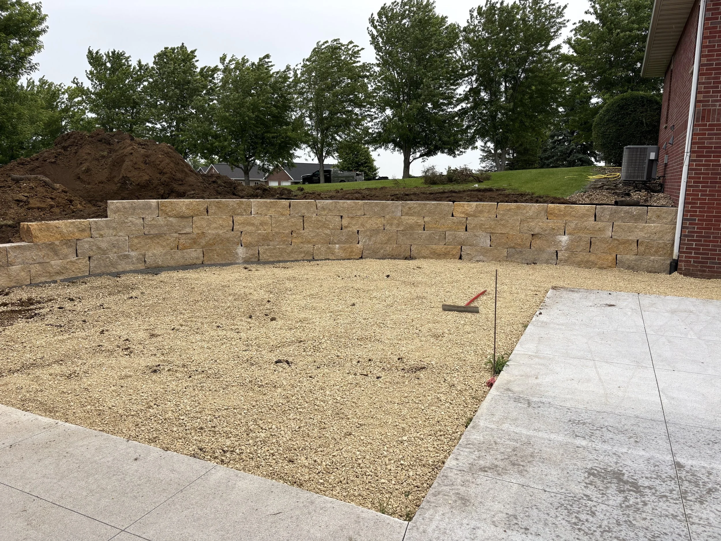 A backyard with a freshly built stone retaining wall, a dirt pile, a concrete patio, and trees in the background, with parts of a brick house on the right and a yellow garden hose coiled near an air conditioning unit.