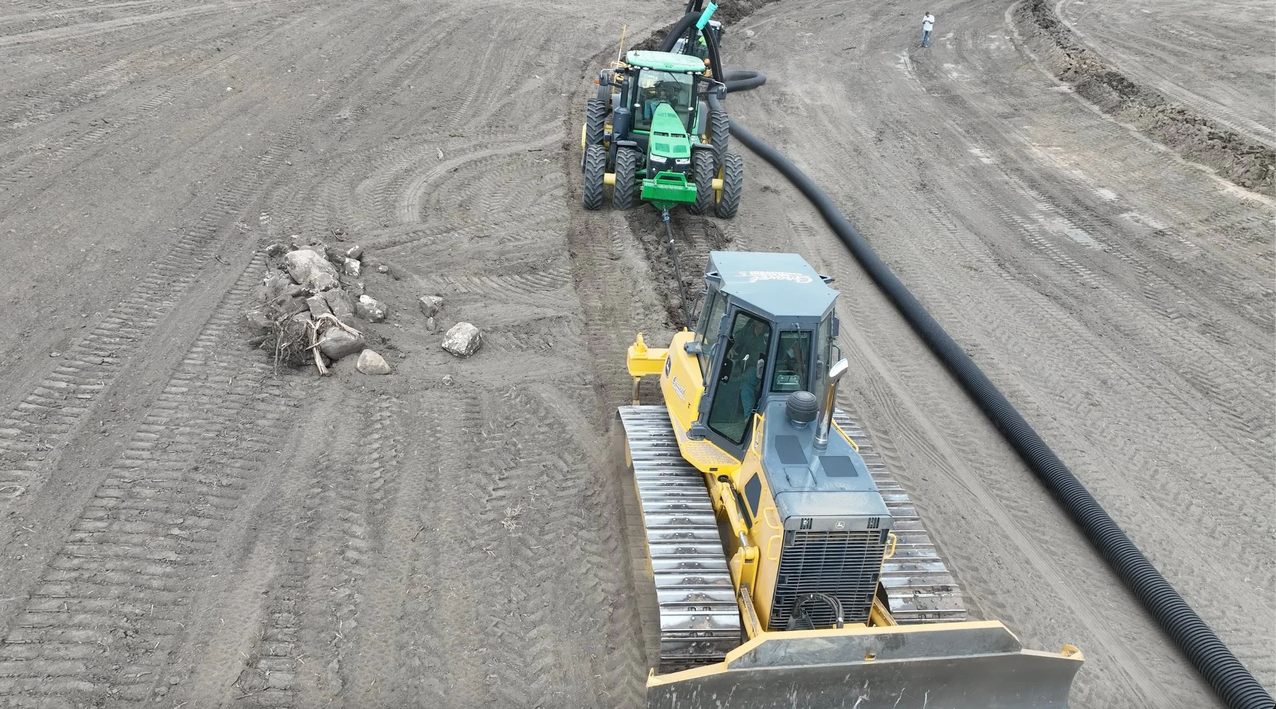 Construction site with a small bulldozer and a large tractor working on leveling the dirt ground. Two workers are standing in the background, and a black pipe is laid along the ground.