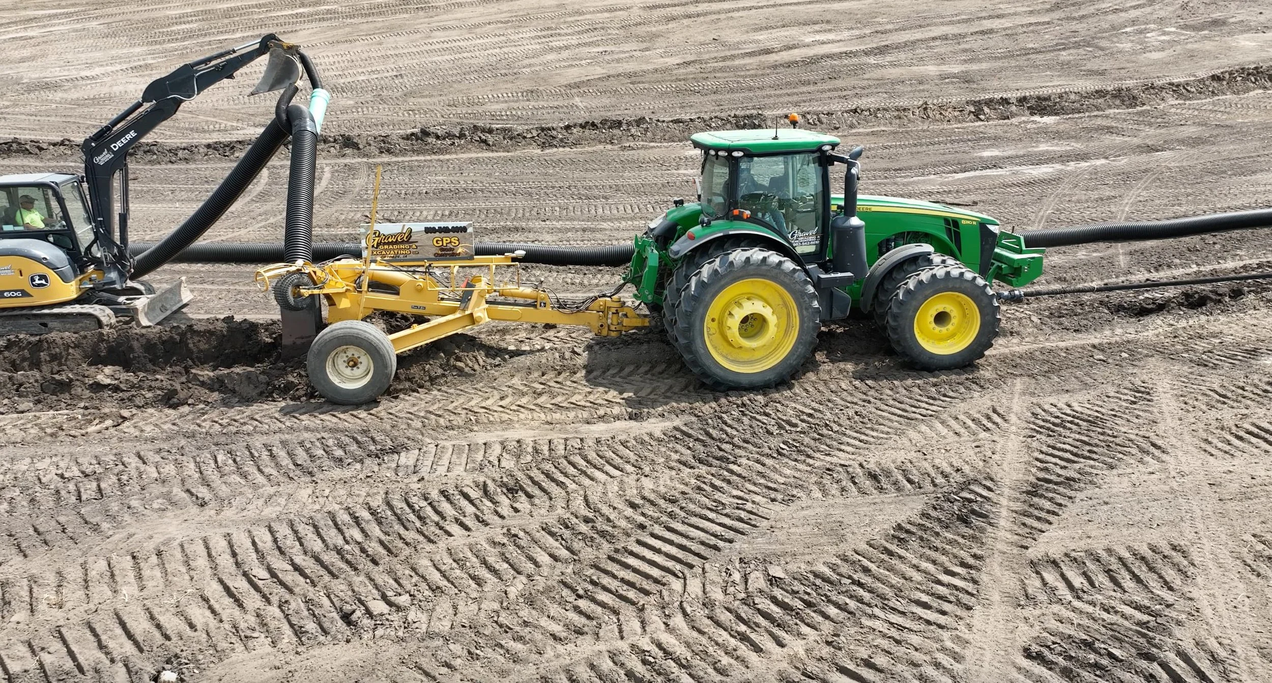 A construction site in an Eastern Iowa farm field and heavy machinery, including a large green John Deere tractor pulling a yellow grading tool, and a small excavator with a corrugated pipe attached.