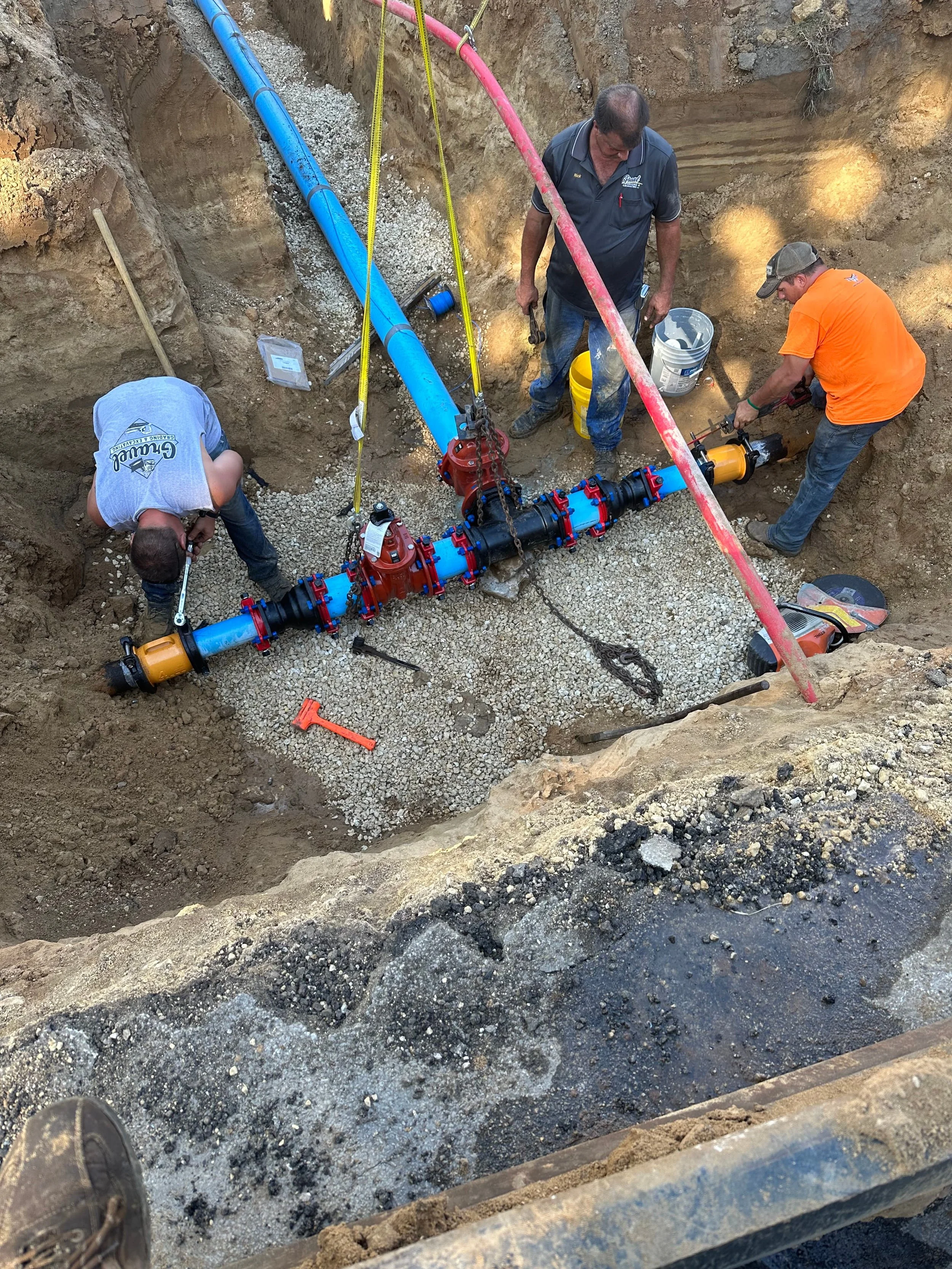 Three workers are repairing a large pipeline in a deep excavation. The pipeline is blue, and the workers are using tools and wearing casual work clothes. One worker is wearing an orange shirt and a baseball cap, another is in a gray t-shirt, and the third is in a dark-colored polo shirt. The area is surrounded by dirt and gravel.