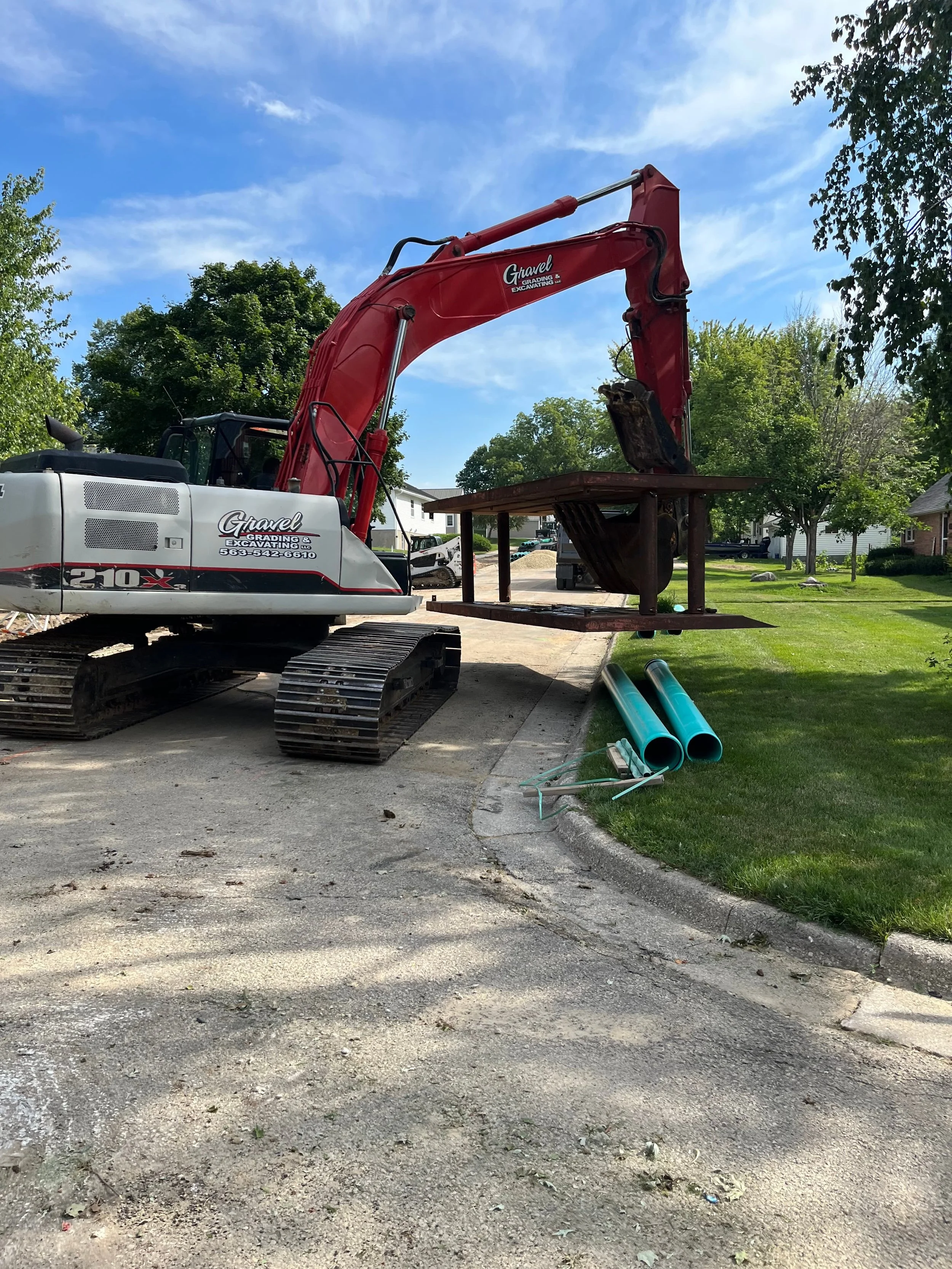 A red and white excavator lifting a metal platform on a residential street with green trees and houses in the background.