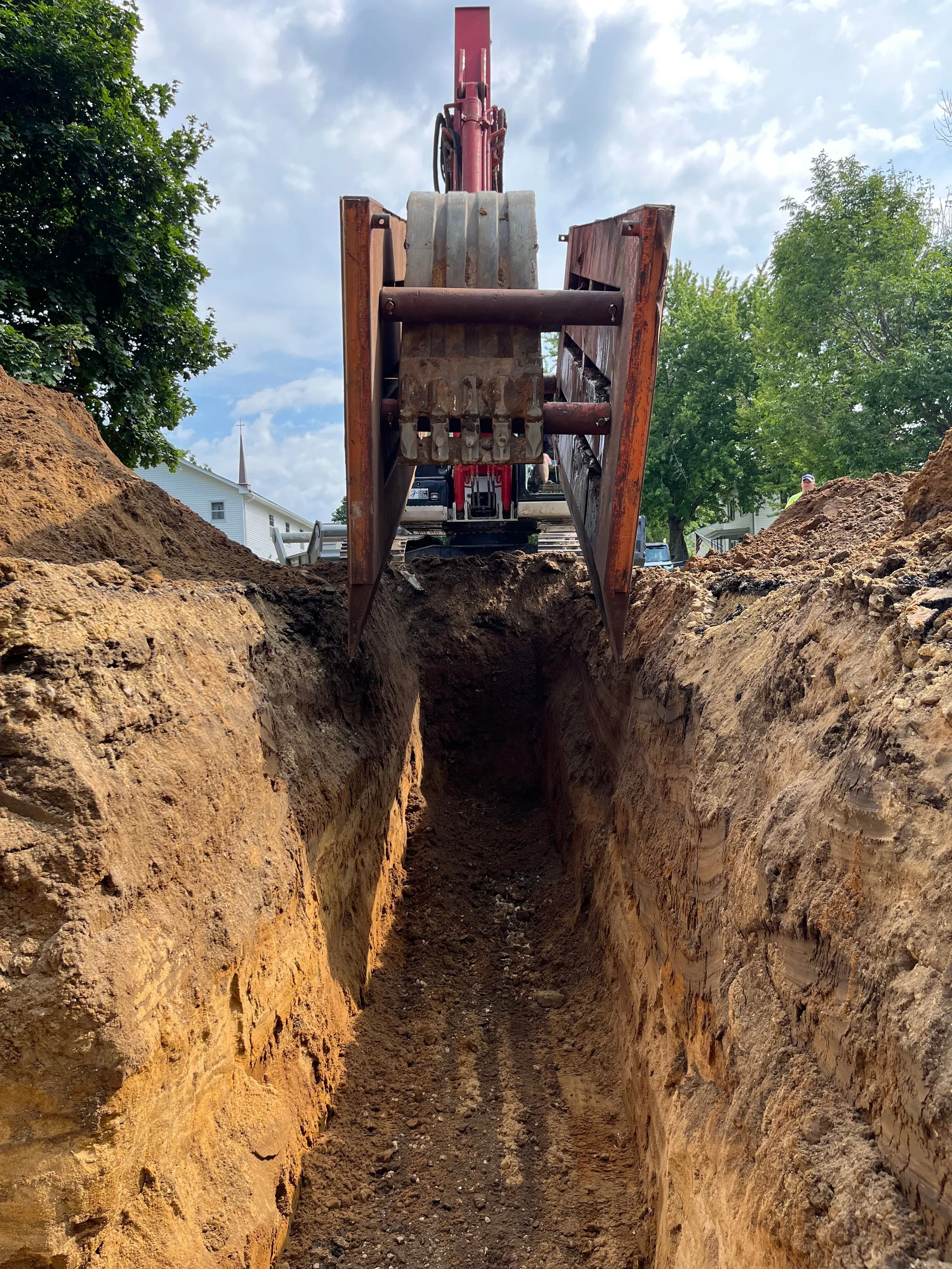 View of a construction site featuring a deep trench with excavated soil on both sides. An excavator is positioned at the top of the trench, ready to continue digging.