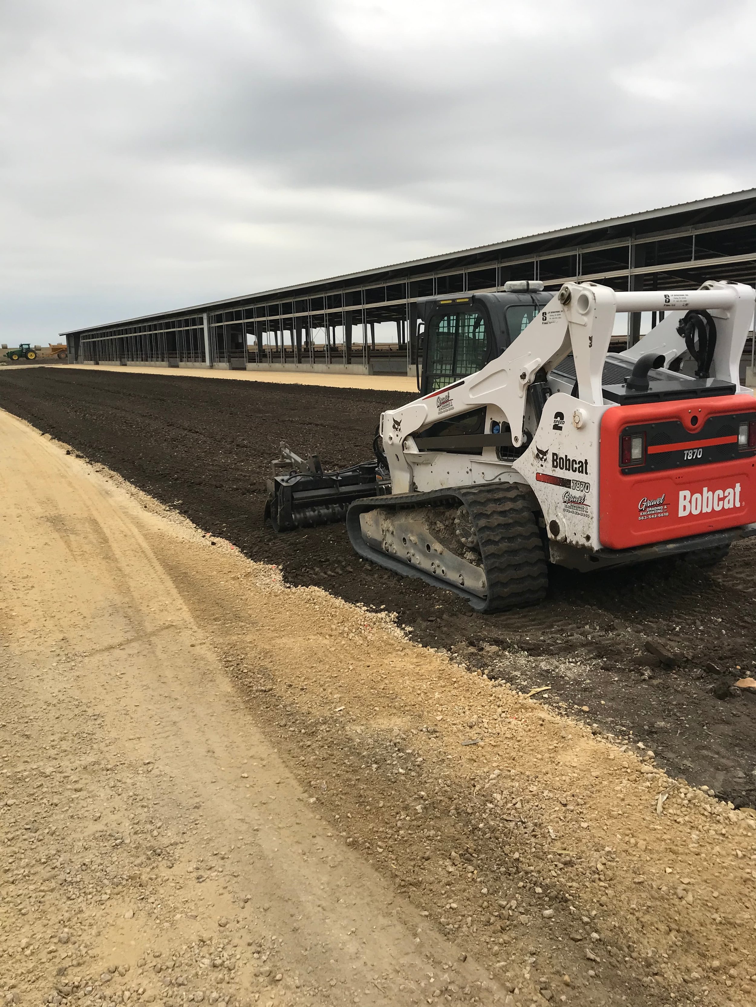 Construction site with a Bobcat skid-steer loader and a long structure under construction, with soil and gravel ground, cloudy sky.