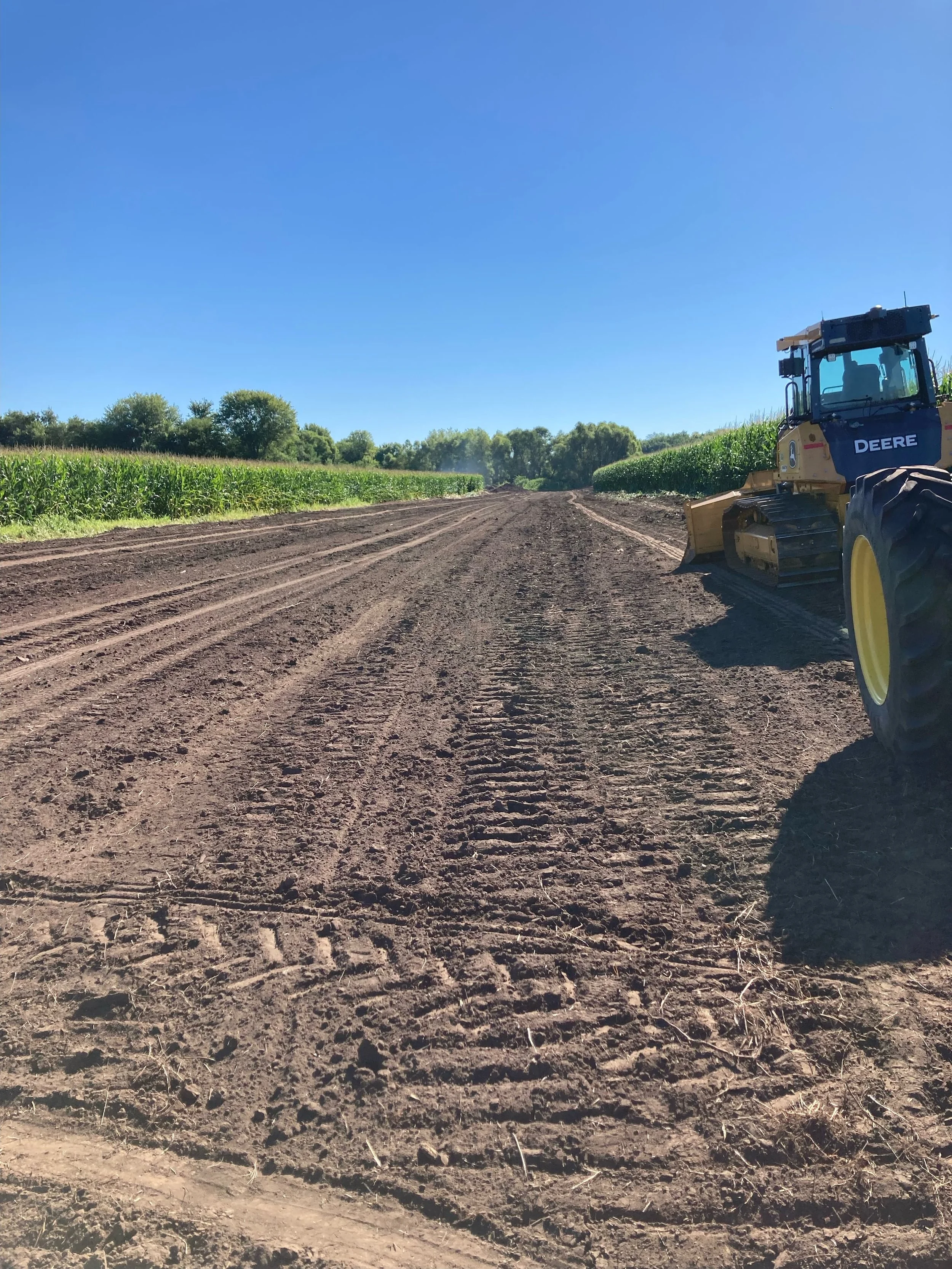 A dirt farm road with a yellow John Deere bulldozer on the right side, green crops on both sides, and a line of trees under a clear blue sky.