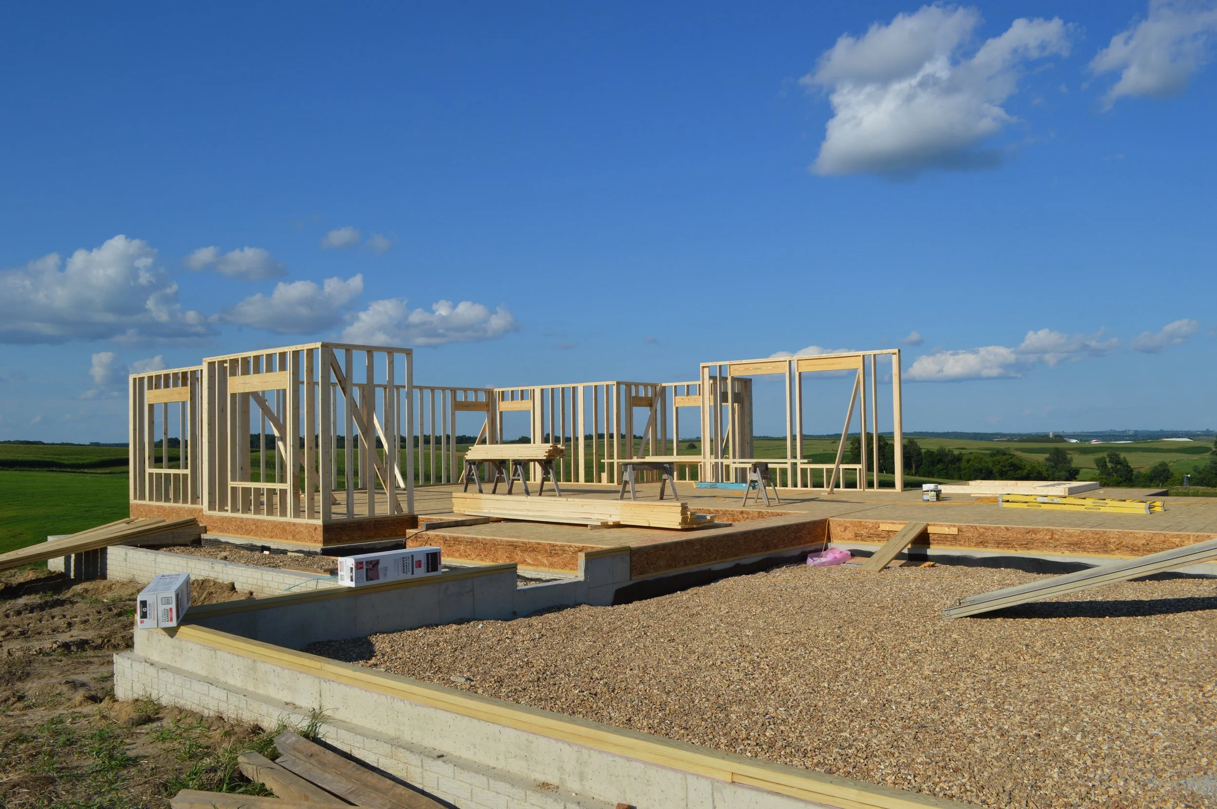 Construction site with wooden framing for a house set against a blue sky with clouds and green fields in the background.
