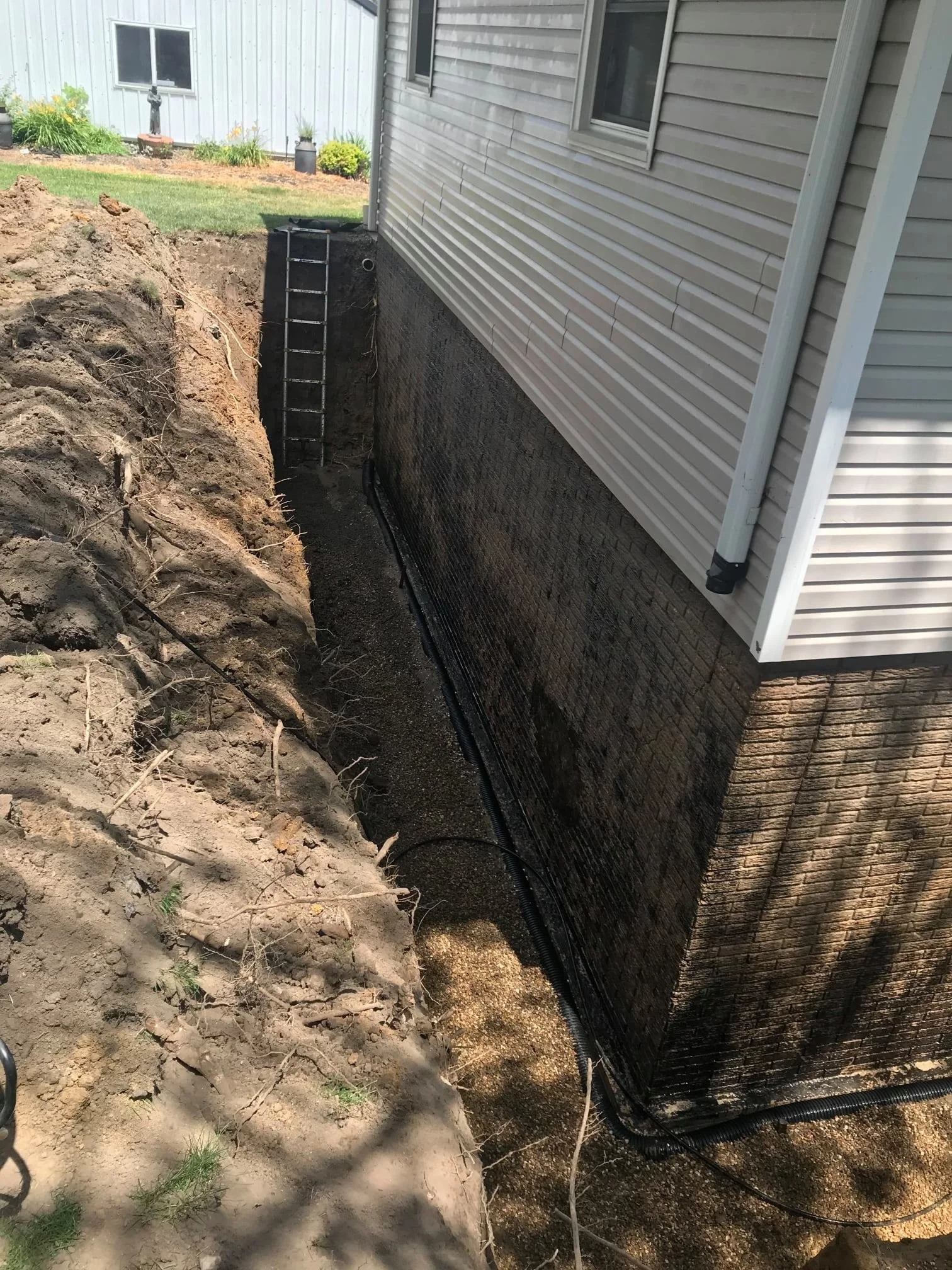Excavation site next to a house, with a trench dug along the foundation wall, a ladder inside the trench, and a black drainage pipe at the bottom. Dirt and some roots are in the foreground, with part of the house's siding and windows visible.