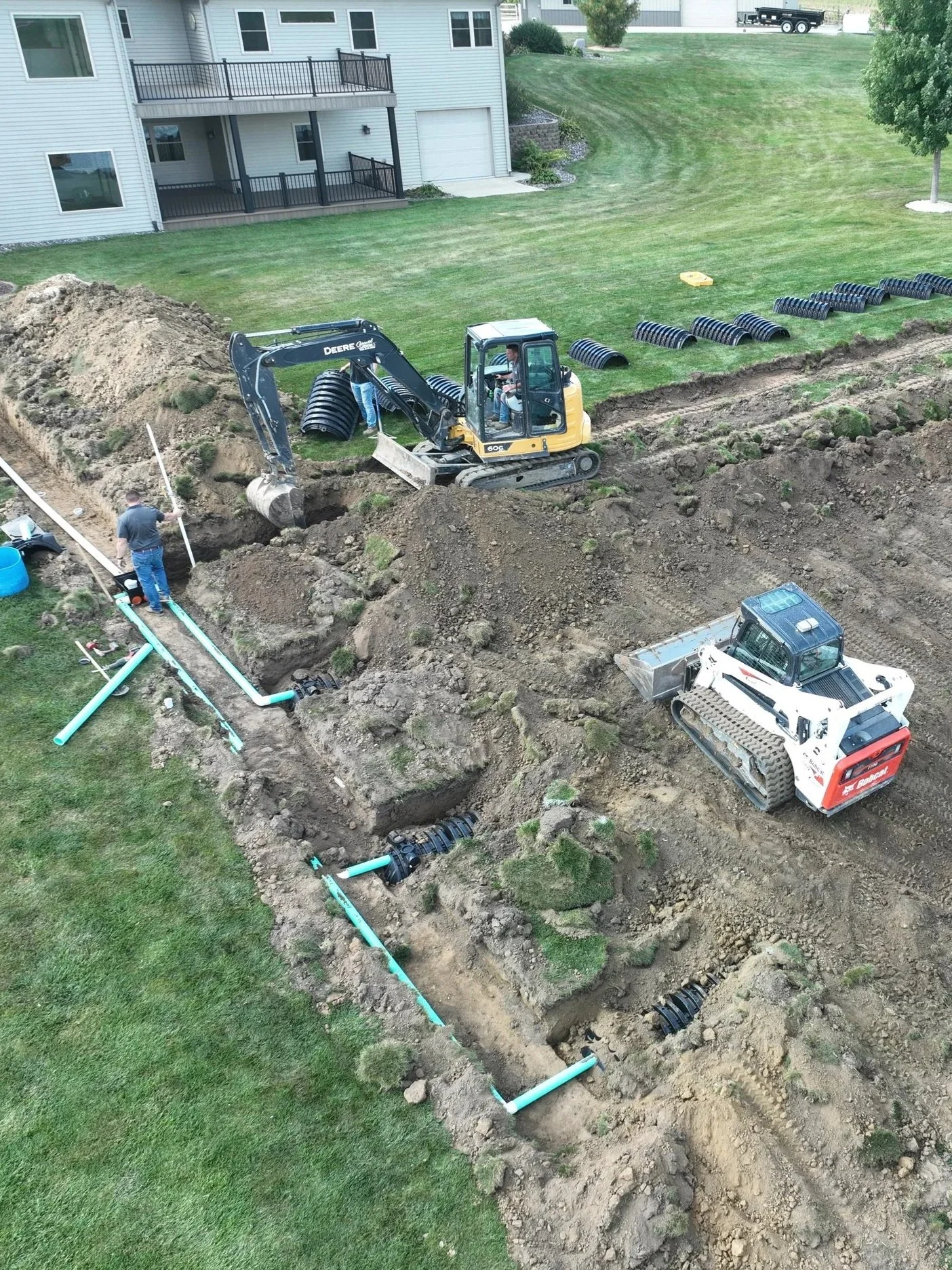 Construction workers installing underground piping for septic system with a small excavator on a grassy backyard next to a white house with a deck.