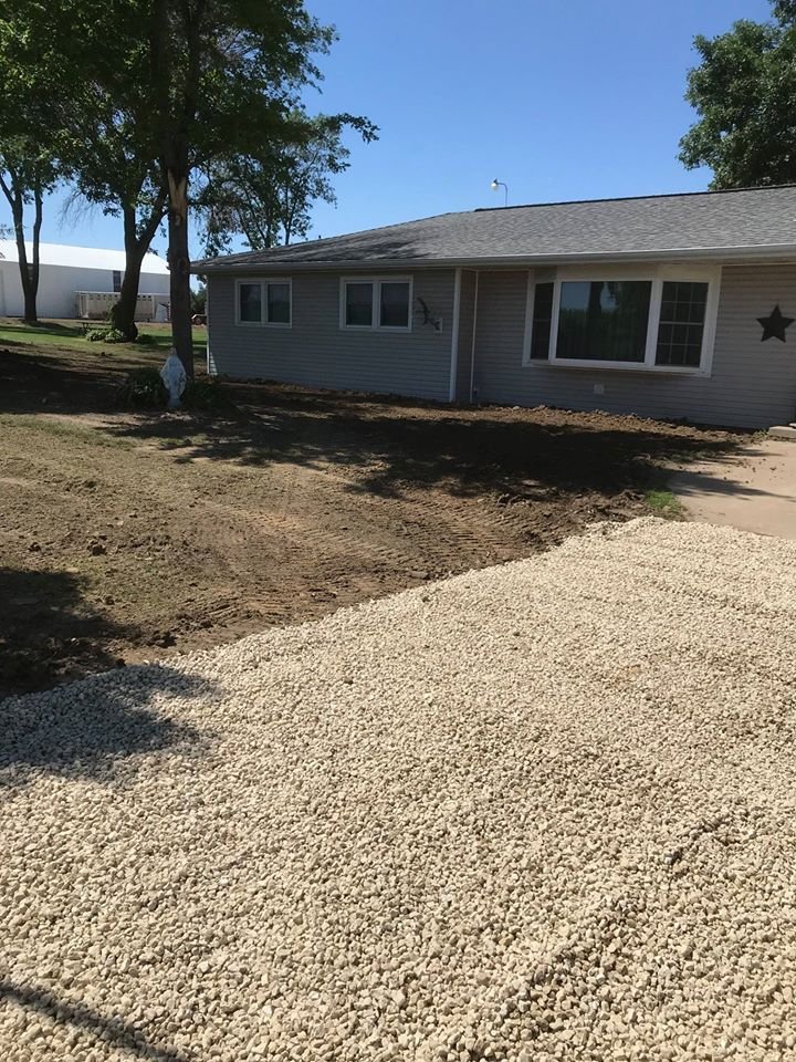 A house with a grey exterior and a black star decoration on the wall, surrounded by trees, with a patch of gravel driveway in the foreground and dirt yard in the background.