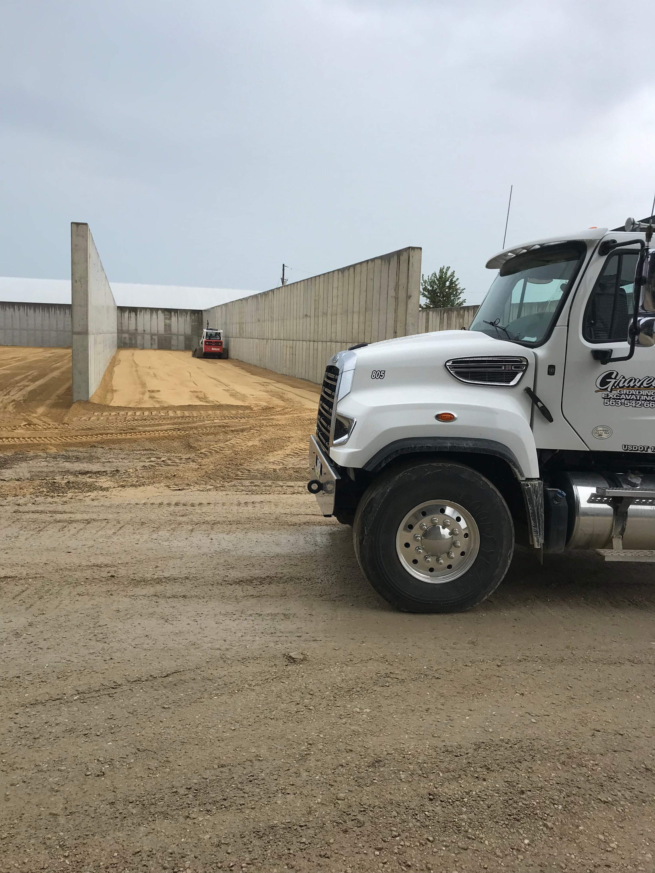 Construction site with a gravel ground, a white truck on the right, and a small skid-steer loader in the background near a concrete barrier at a feed bunk site in Eastern Iowa.