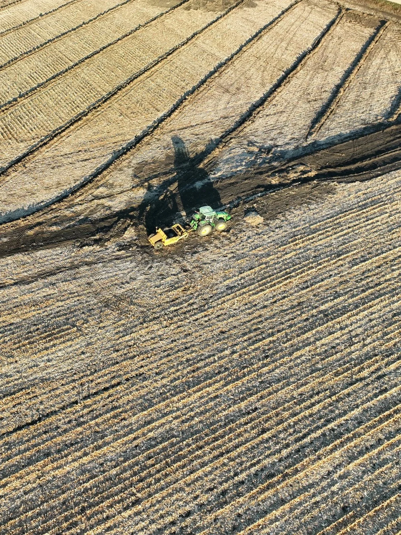 An aerial view of farmland with pattern tile being installed by a green tractor pulling a yellow equipment, creating a large shadow on the ground.