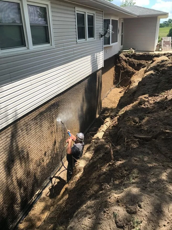 A worker power washing the foundation of a house from a trench dug along the side of the house, with dirt mounds and construction materials visible nearby.