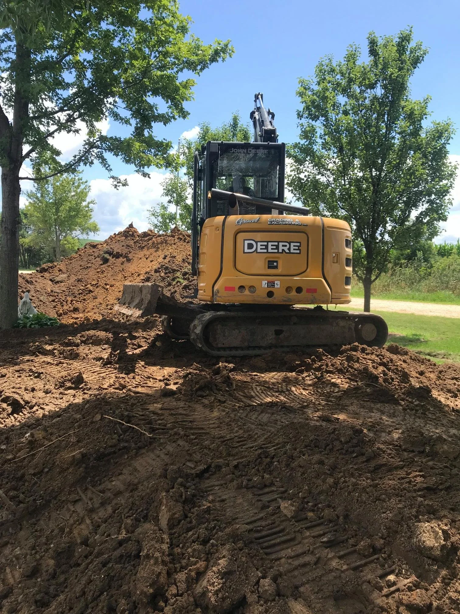 A yellow Deere excavator working on a construction site, digging up dirt surrounded by trees and a dirt ground under a clear blue sky.