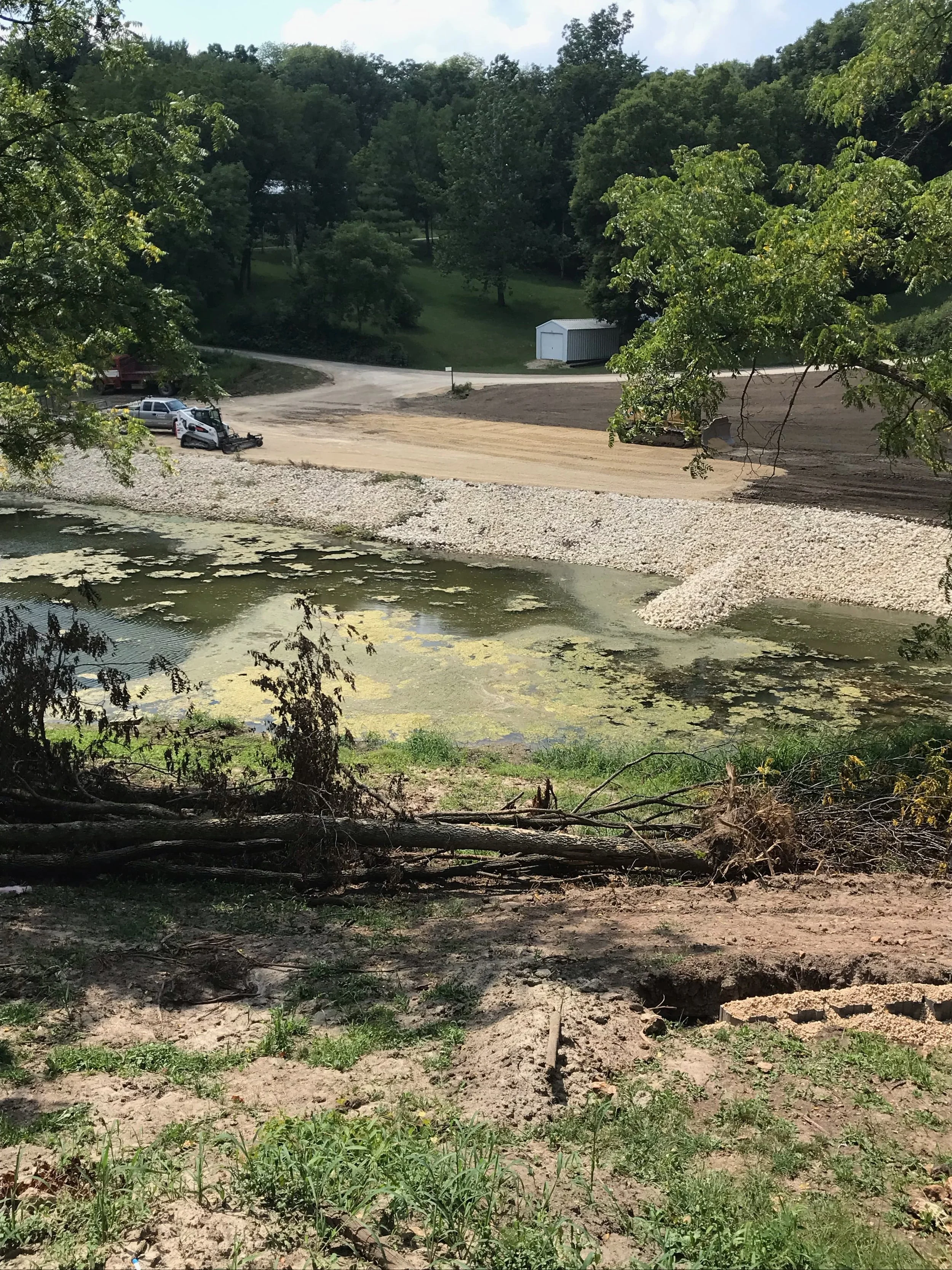 A small dirt path runs through a grassy area next to a body of water with algae. In the background, there is a white storage shed and a wooded hill. Construction equipment and vehicles are parked near the path, indicating some construction or landscaping work.